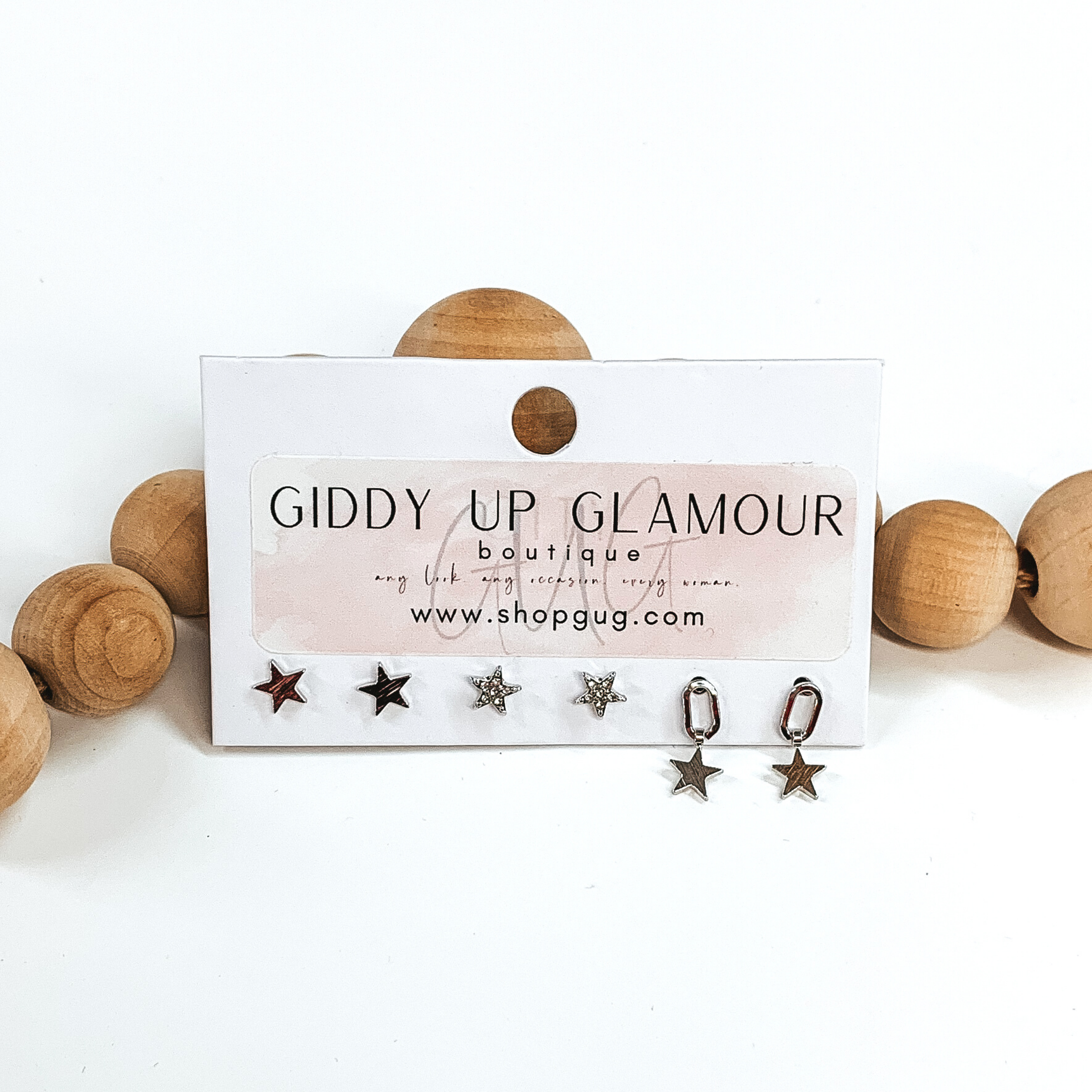 This is a 3 piece silver earring set. There are two sets of silver stud earrings; one is plain and the other has clear crystals. The last earrings is an open oval with a hanging star charm. These earrings are pictured on a Giddy Up Glamour earring holder laying in front of tan beads on a white background