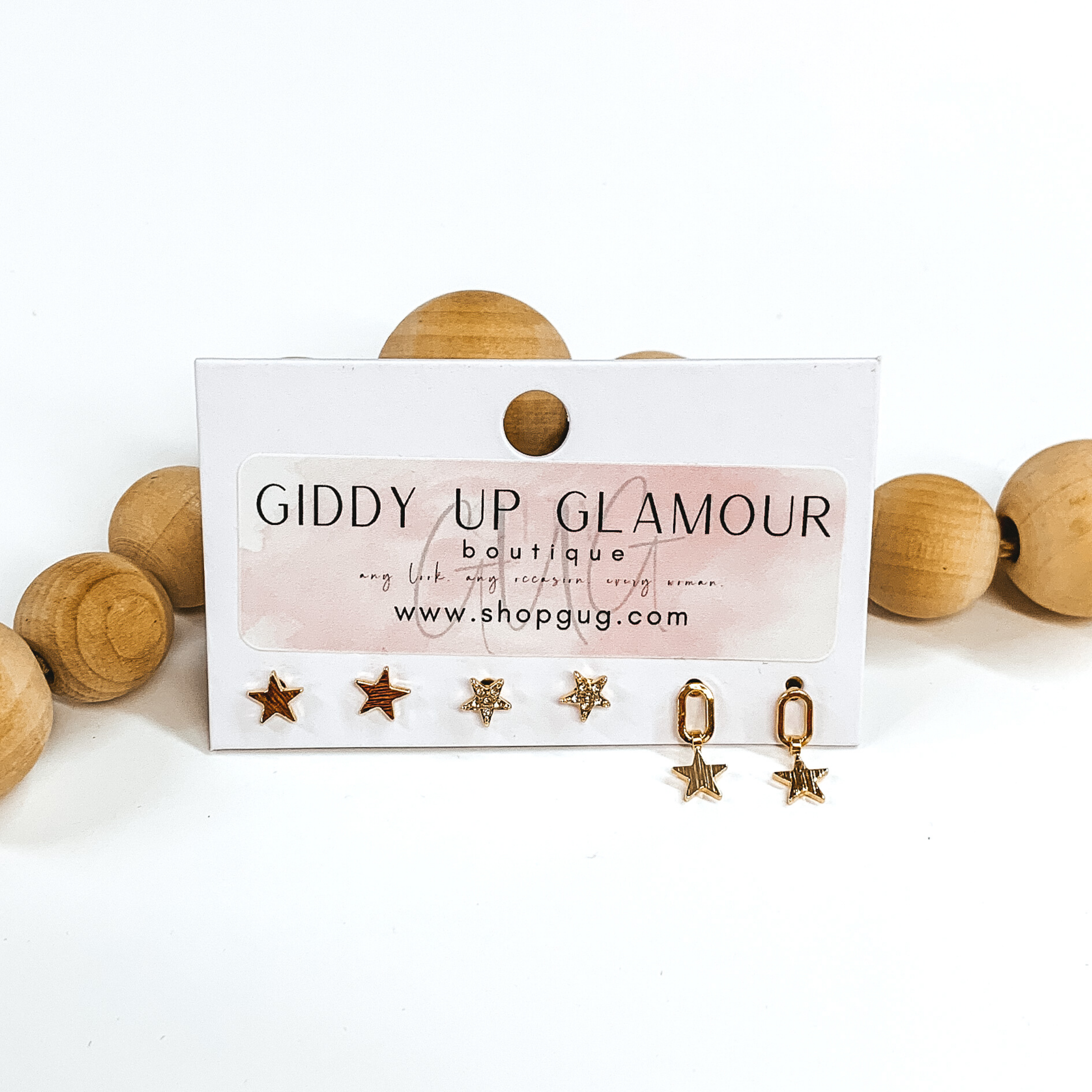 This is a 3 piece gold earring set. There are two sets of gold stud earrings; one is plain and the other has clear crystals. The last earrings is an open oval with a hanging star charm. These earrings are pictured on a Giddy Up Glamour earring holder laying in front of tan beads on a white background.