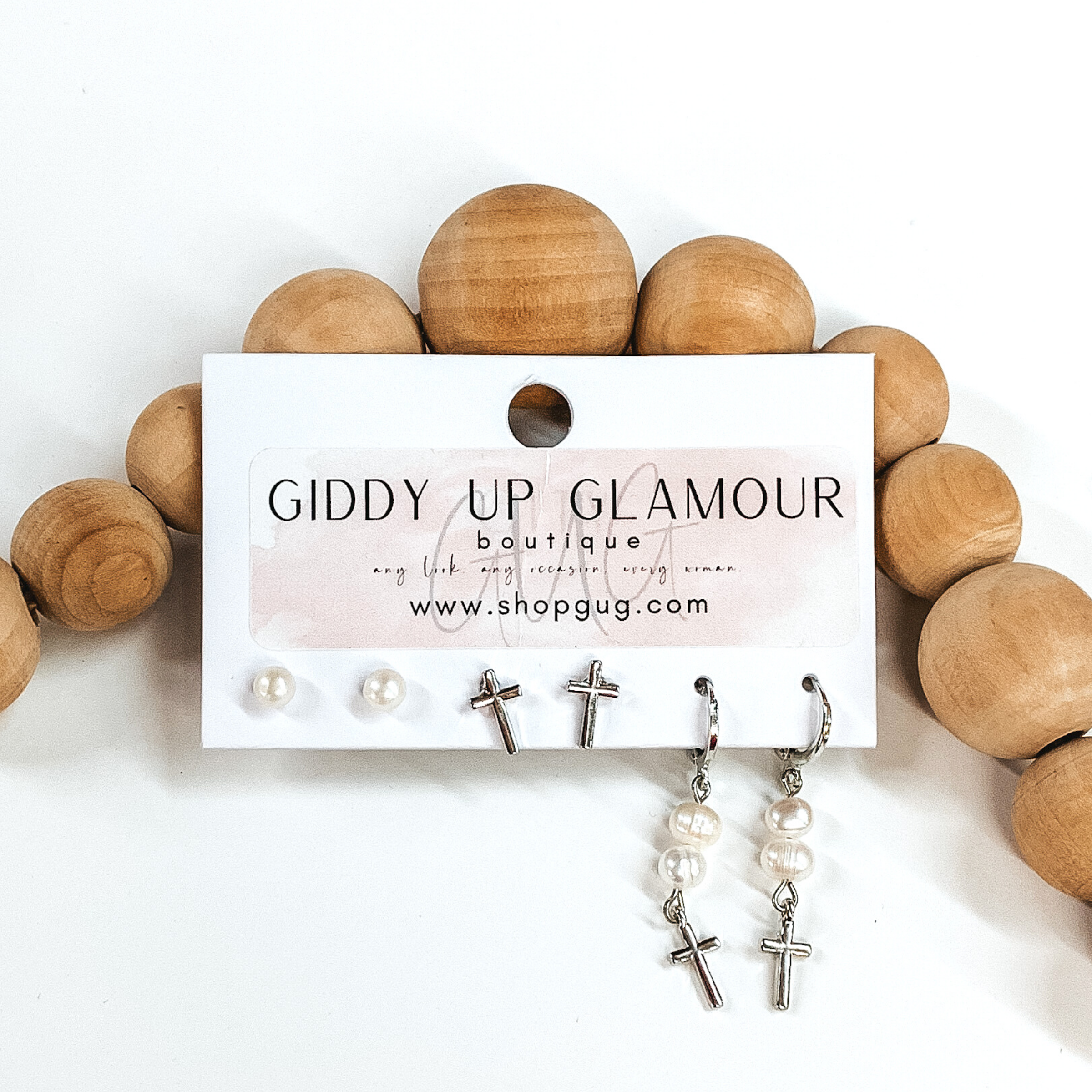 This is a 3 piece silver and pearl earring set. There is a pair of white pearl studs, a silver pair of silver studs, and a pair of dangle pearls with hanging cross pendanta. These earrings are pictured on a Giddy Up Glamour earring holder laying in front of tan beads on a white background. 