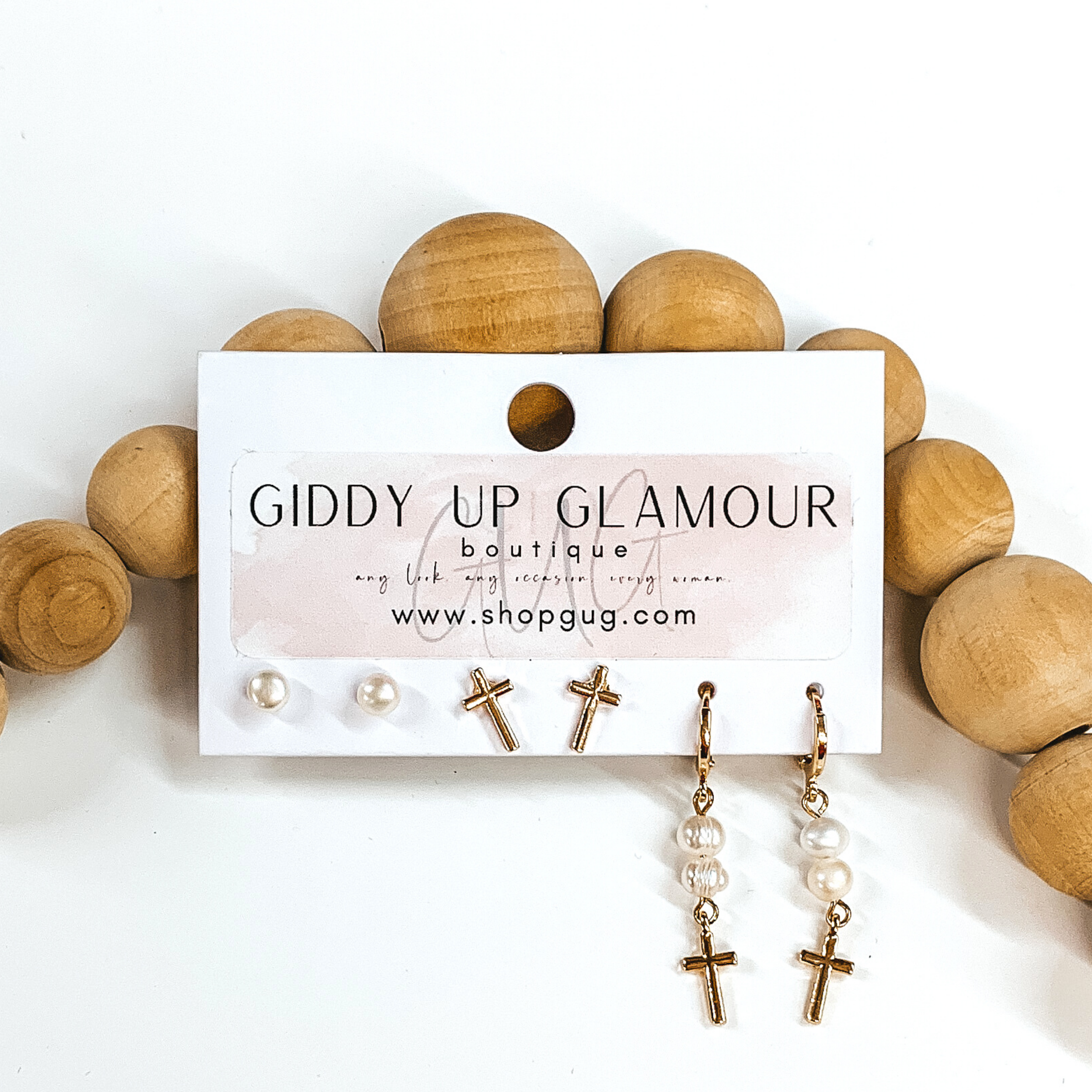 This is a 3 piece gold and pearl earring set. There is a pair of white pearl studs, a gold pair of silver studs, and a pair of dangle pearls with hanging cross pendant. These earrings are pictured on a Giddy Up Glamour earring holder laying in front of tan beads on a white background.  