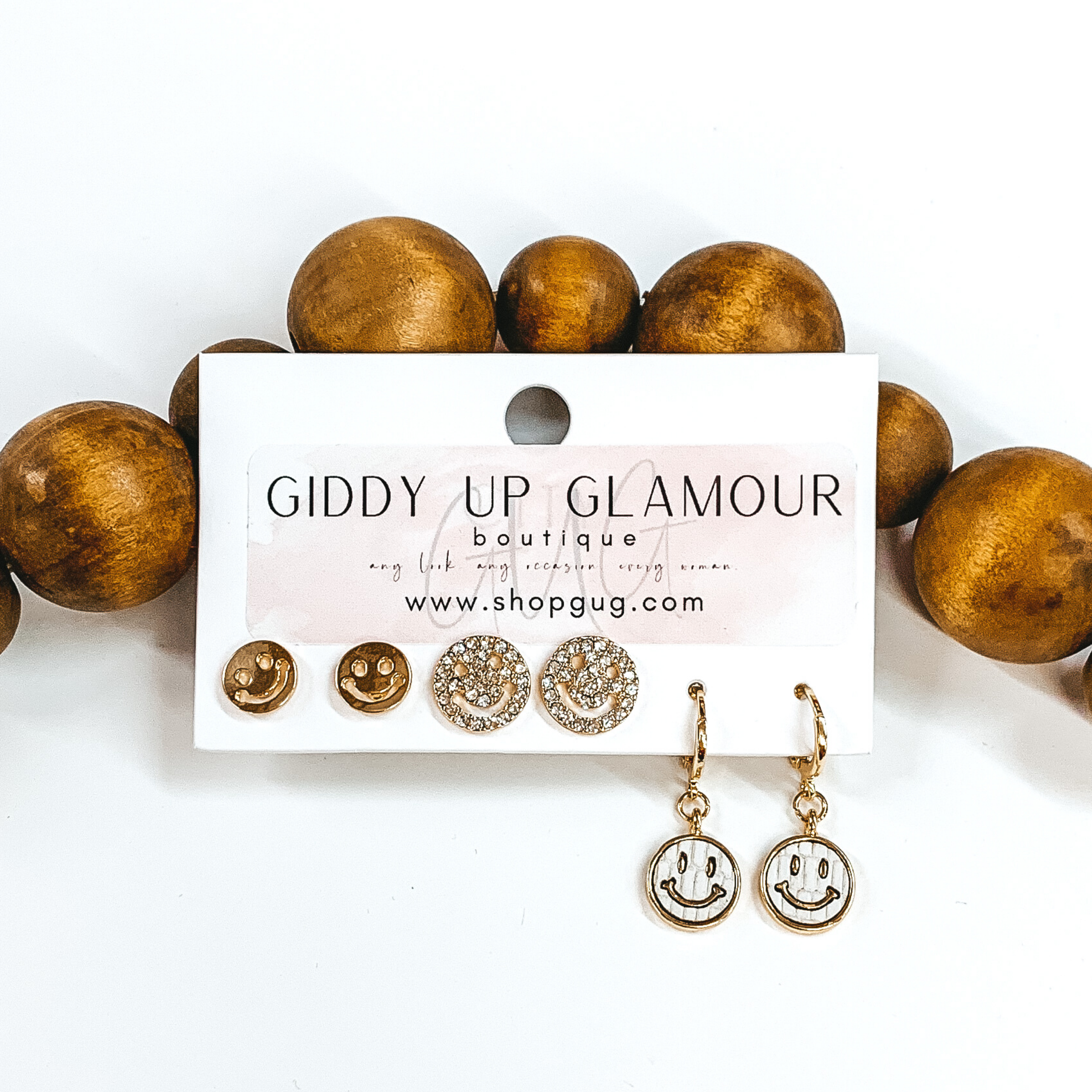 This is a 3 piece gold and pearl earring set. There is a pair of gold smiley face studs, a gold pair smiley face studs with clear crystals, and a pair of dangle pearls with hanging circle white pendant with a gold smiley face. These earrings are pictured on a Giddy Up Glamour earring holder laying in front of tan beads on a white background.