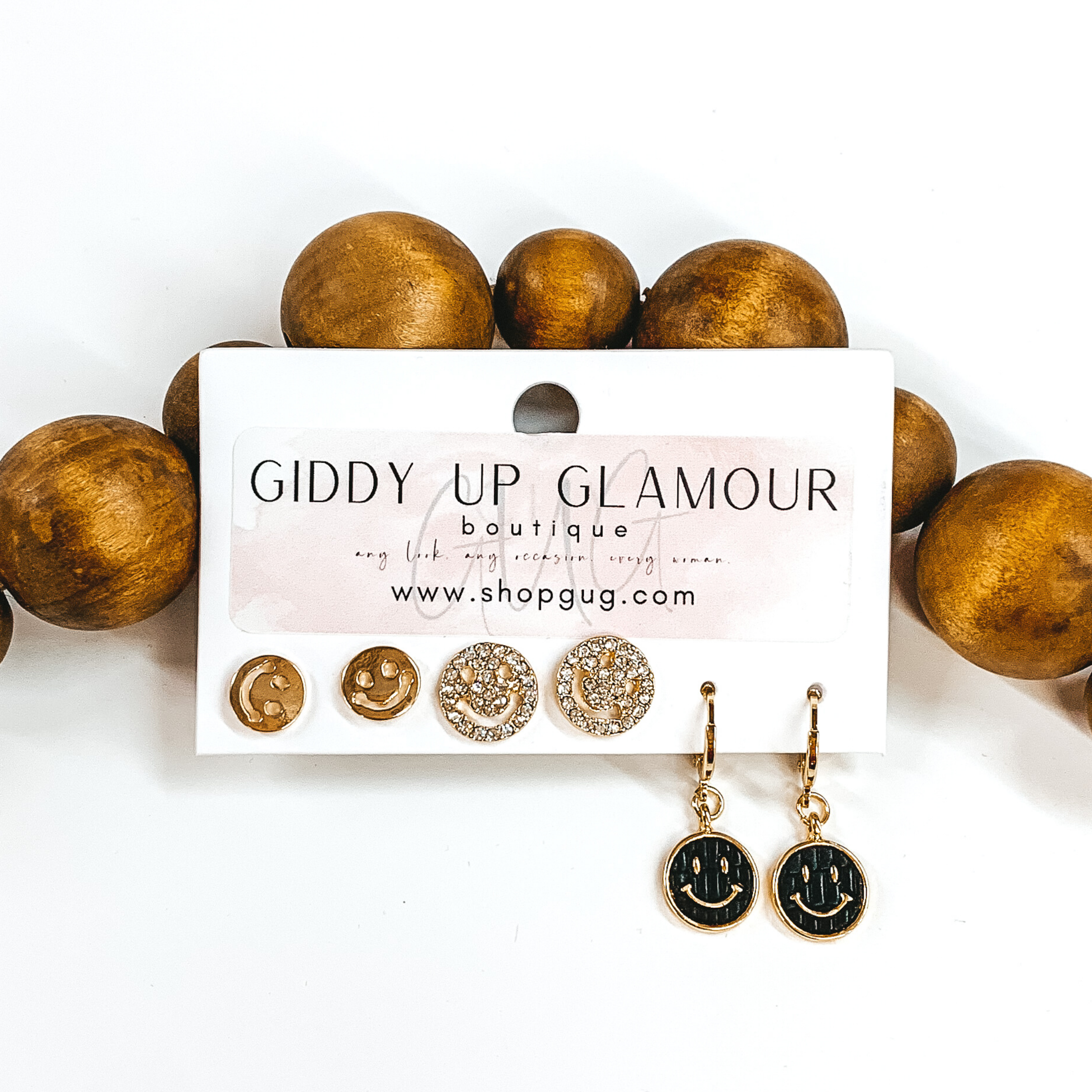 This is a 3 piece gold and pearl earring set. There is a pair of gold smiley face studs, a gold pair smiley face studs with clear crystals, and a pair of dangle pearls with hanging circle black pendant with a gold smiley face. These earrings are pictured on a Giddy Up Glamour earring holder laying in front of tan beads on a white background.  