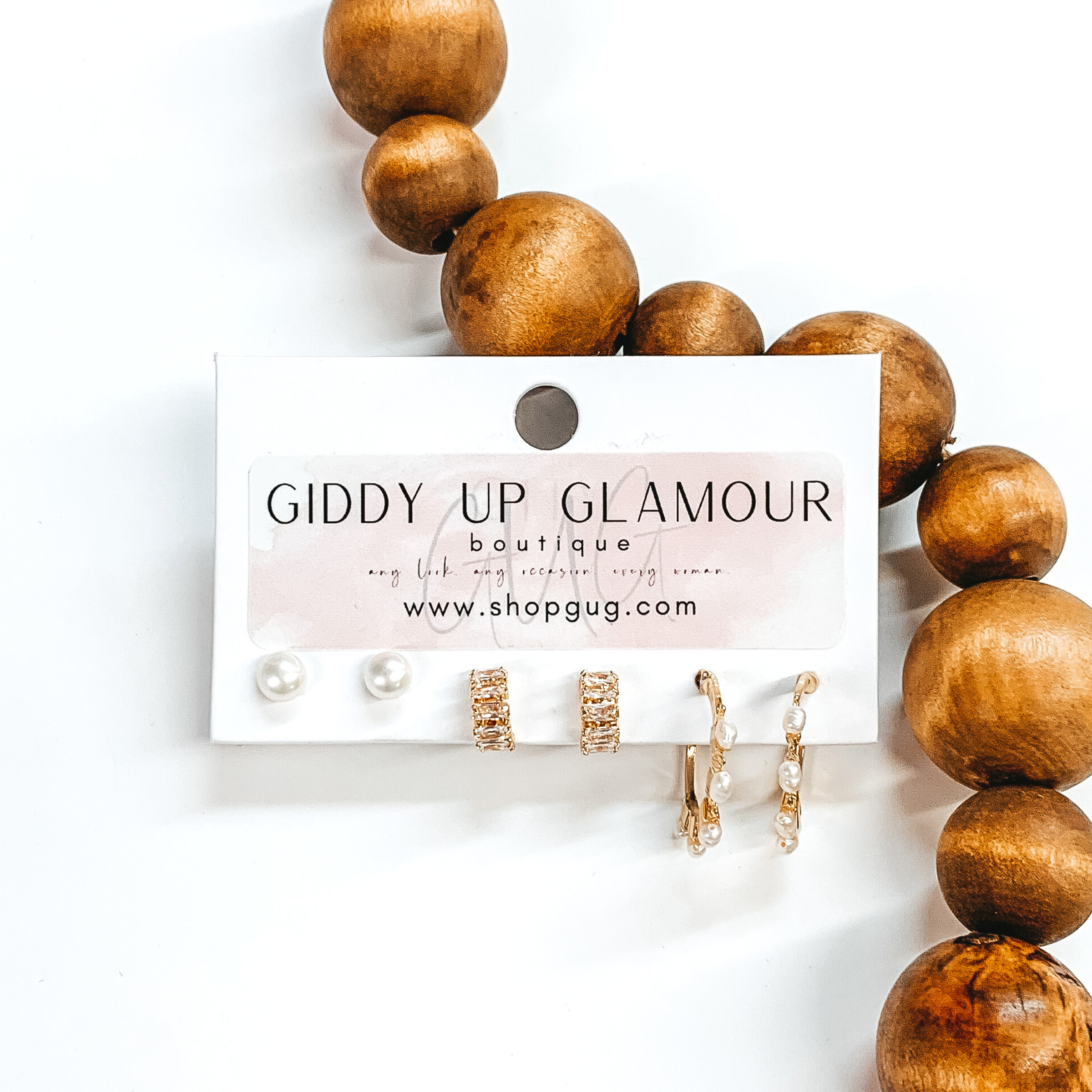 This is a 3 piece crystal and pearl gold earring set. There is a pair of white pearl studs, a gold pair of tiny half hoops with clear crystal baguettes, and a gold pair of hoops with white pearls. These earrings are pictured on a Giddy Up Glamour earring holder laying in front of brow beads on a white background.