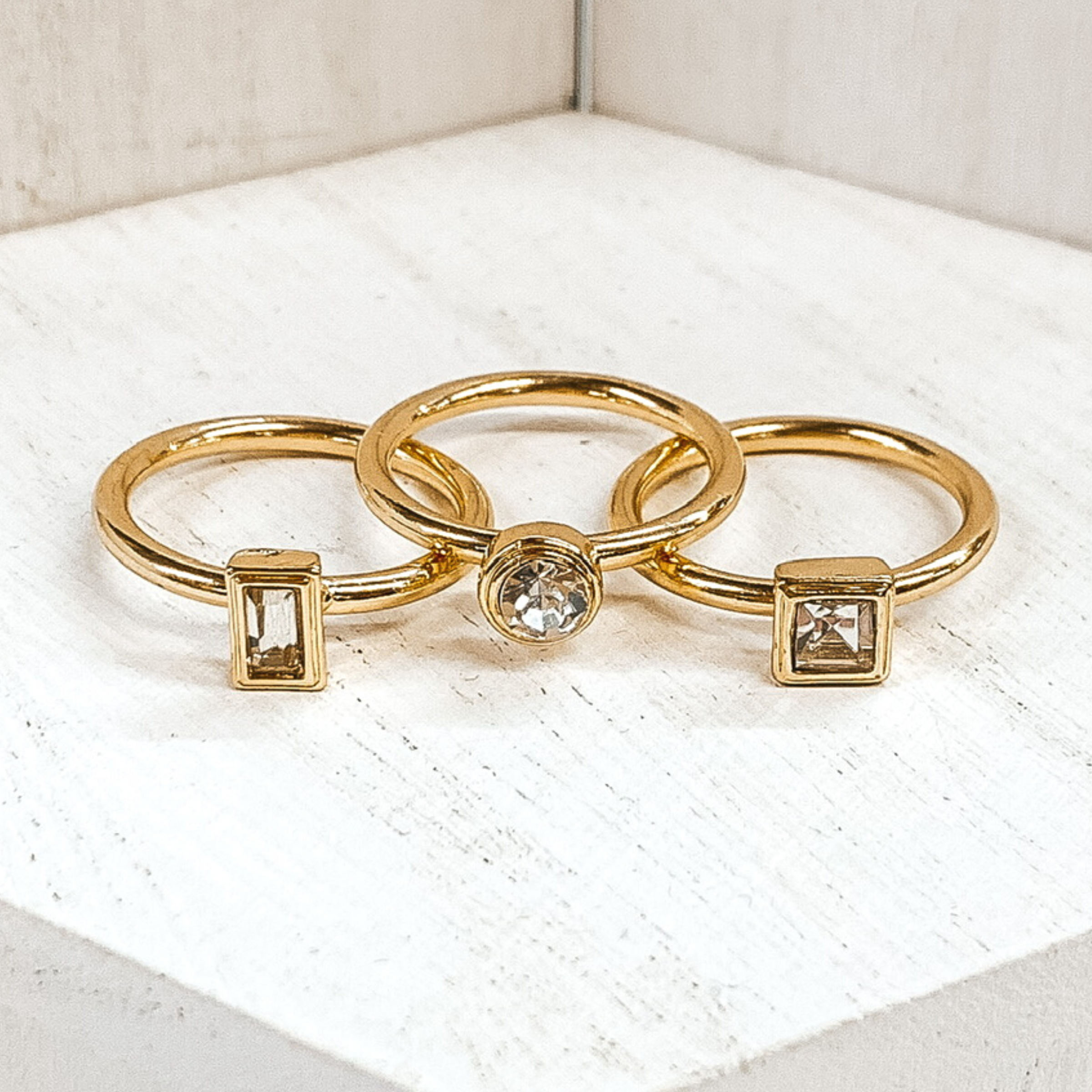 Set of three gold rings. One ring has a small rectangle clear crystal. Another ring has a circle clear crystal. The third ring has a square clear crystal. These rings are pictured on a white background. 