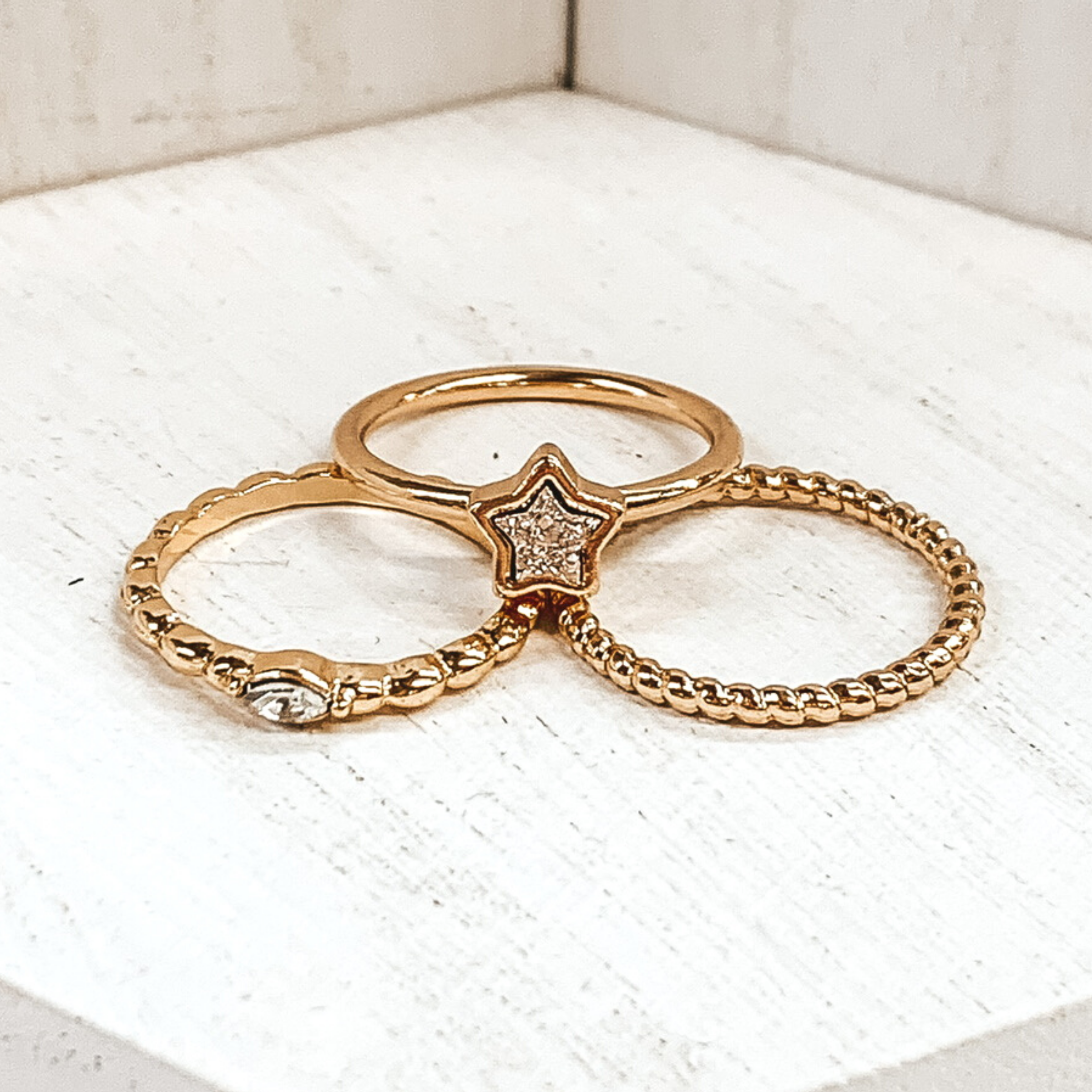 Set of three gold rings. One ring is a twisted texture. Another ring is a bubbled texture with a center oval crystal. The last ring has a plain gold band with a druzy star pendant outlined in gold. These rings are pictured on a white background. 