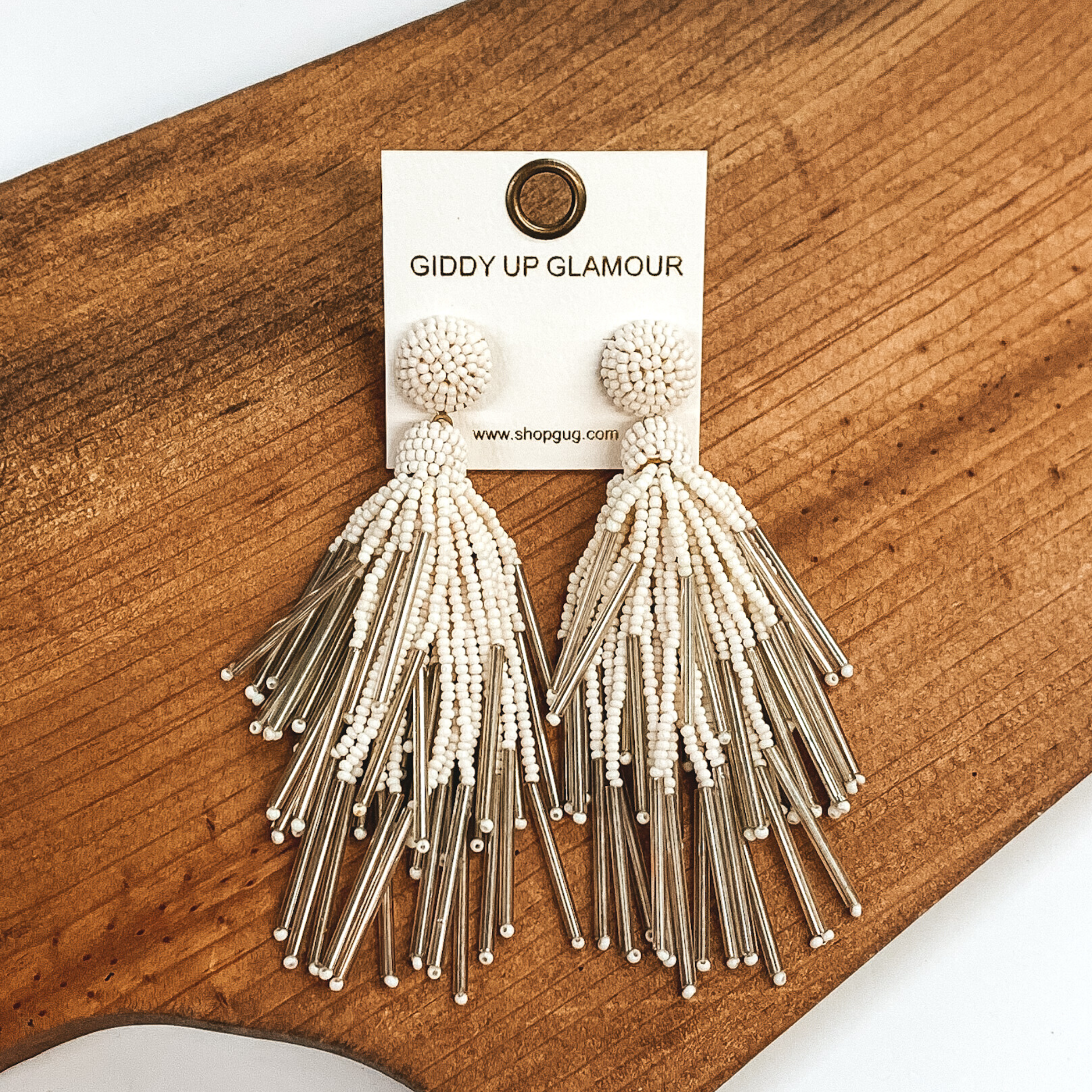 Circle beaded stud earrings with a beaded tassel in ivory. These earrings are pictured laying on a brown piece of wood on a white background.