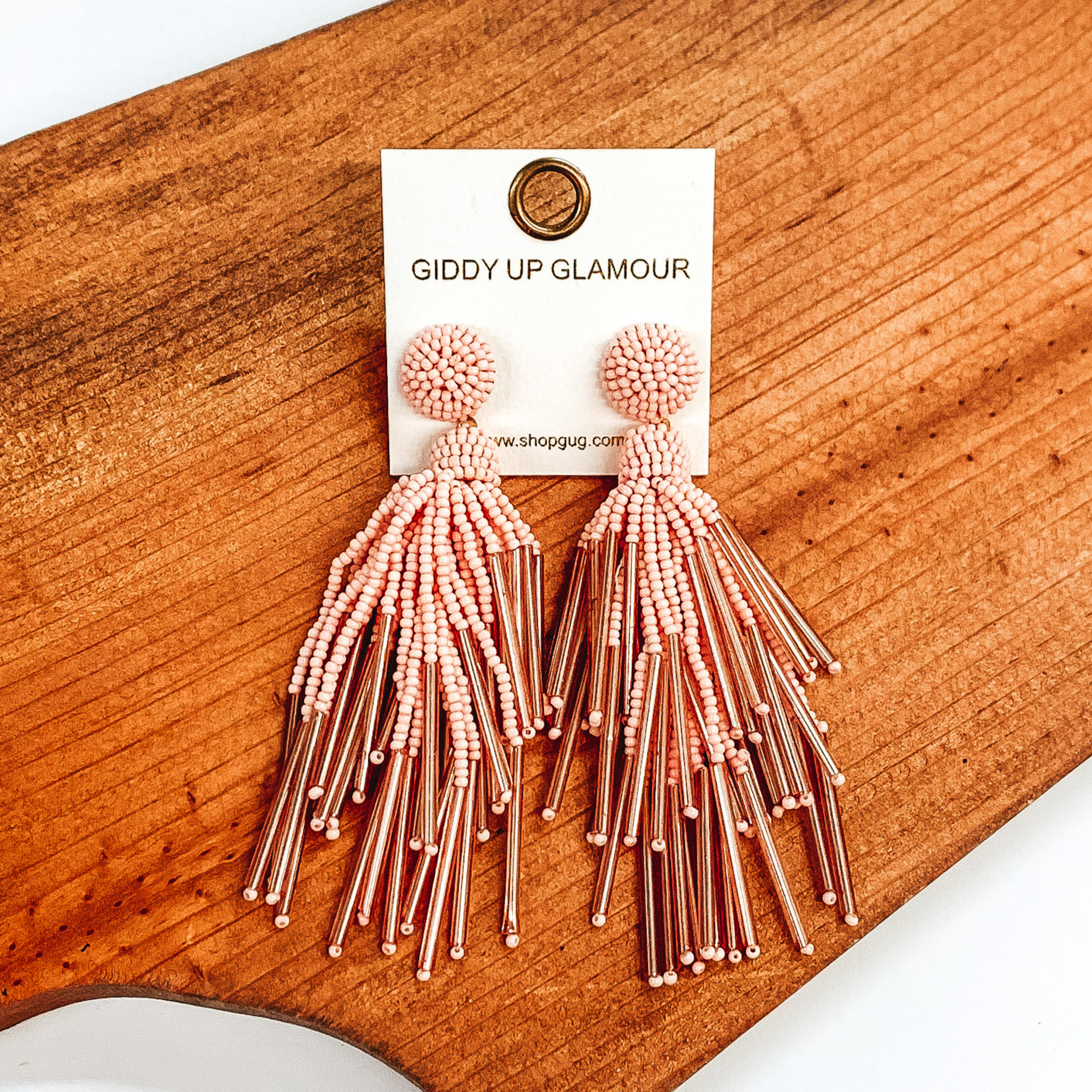 Circle beaded stud earrings with a beaded tassel in baby pink. These earrings are pictured laying on a brown piece of wood on a white background.
