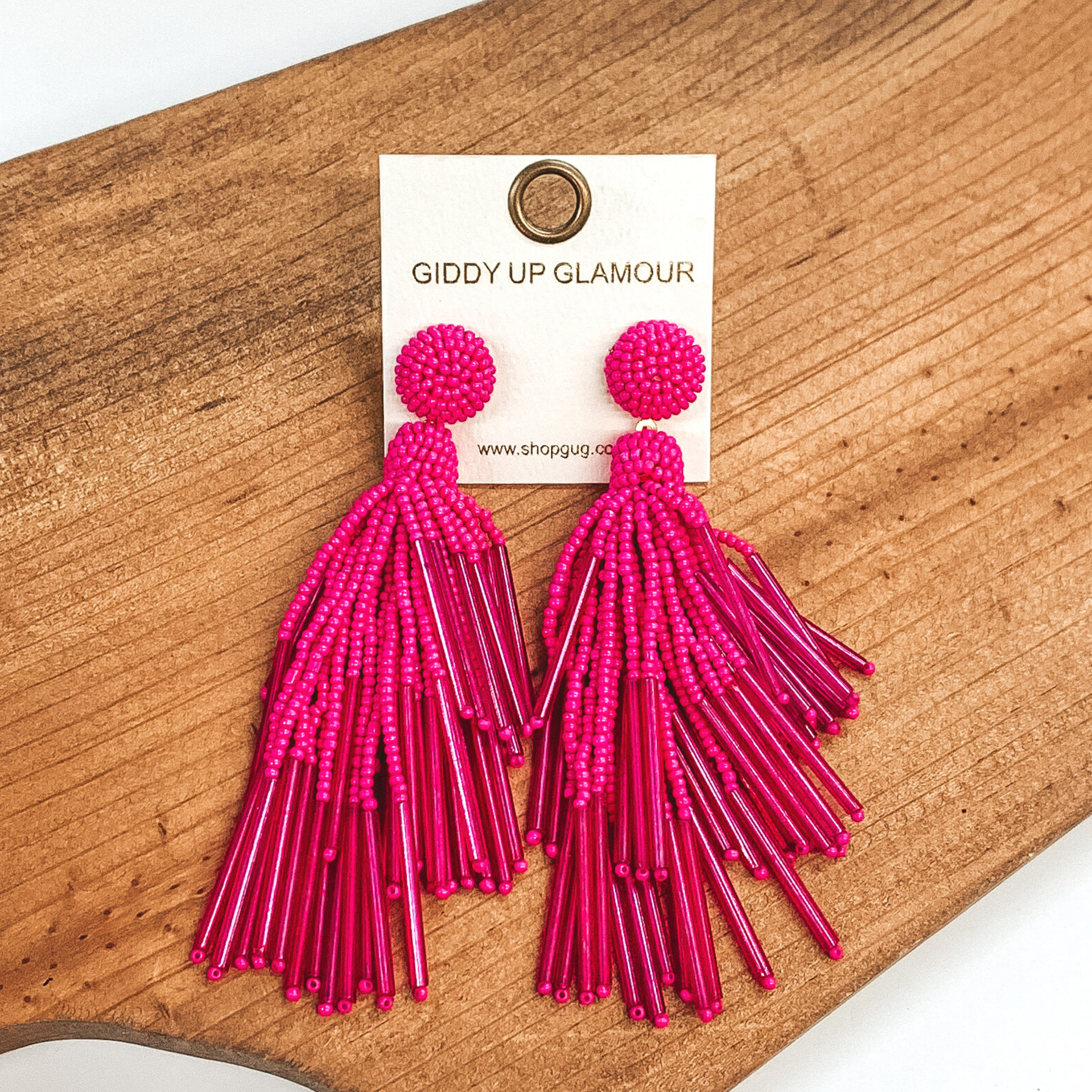Circle beaded stud earrings with a beaded tassel in hot pink. These earrings are pictured laying on a brown piece of wood on a white background.