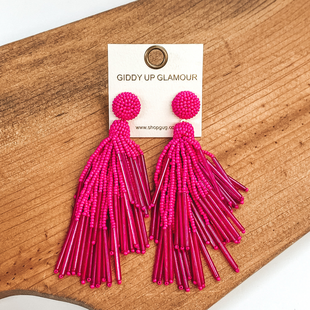 Circle beaded stud earrings with a beaded tassel in hot pink. These earrings are pictured laying on a brown piece of wood on a white background.