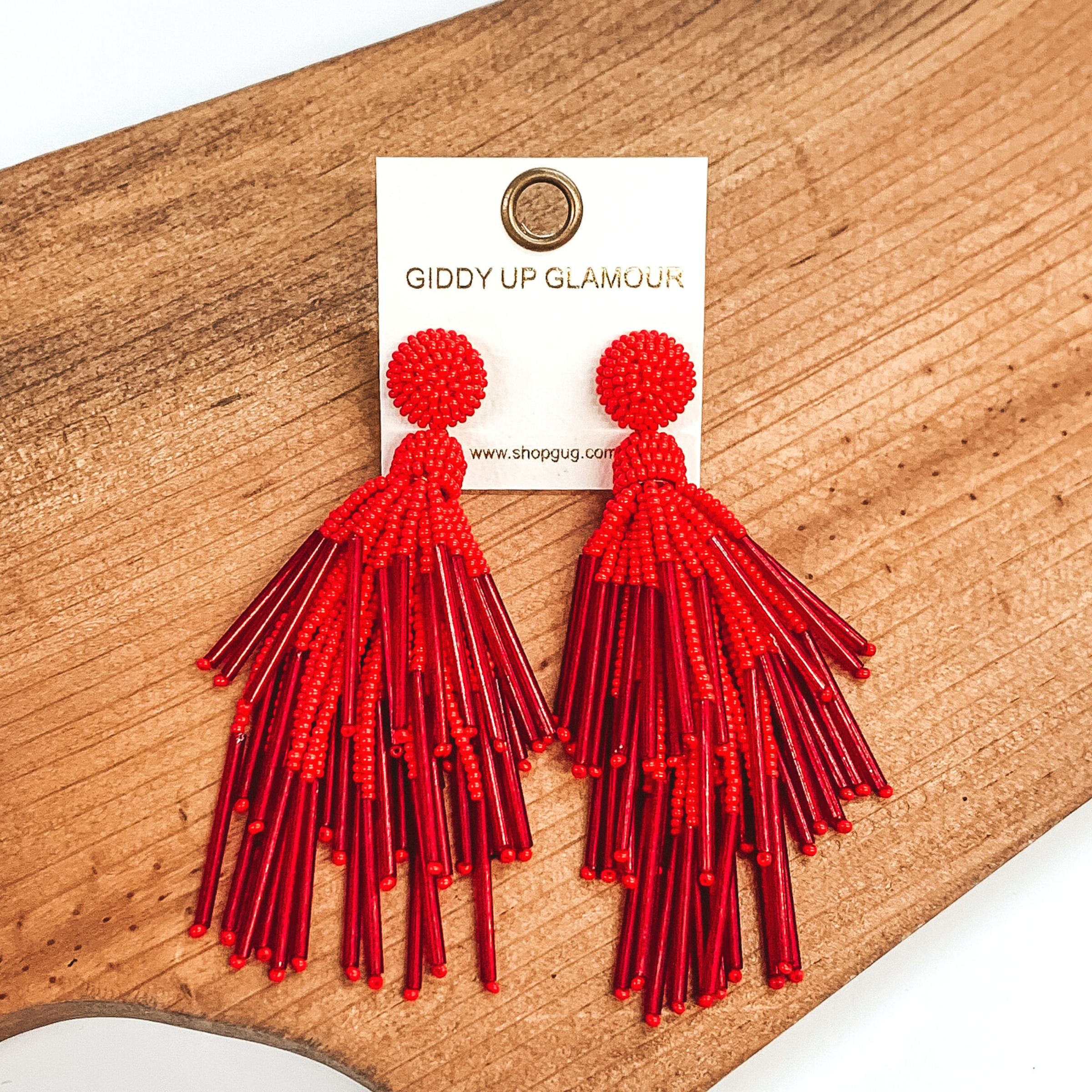 Circle beaded stud earrings with a beaded tassel in red. These earrings are pictured laying on a brown piece of wood on a white background.