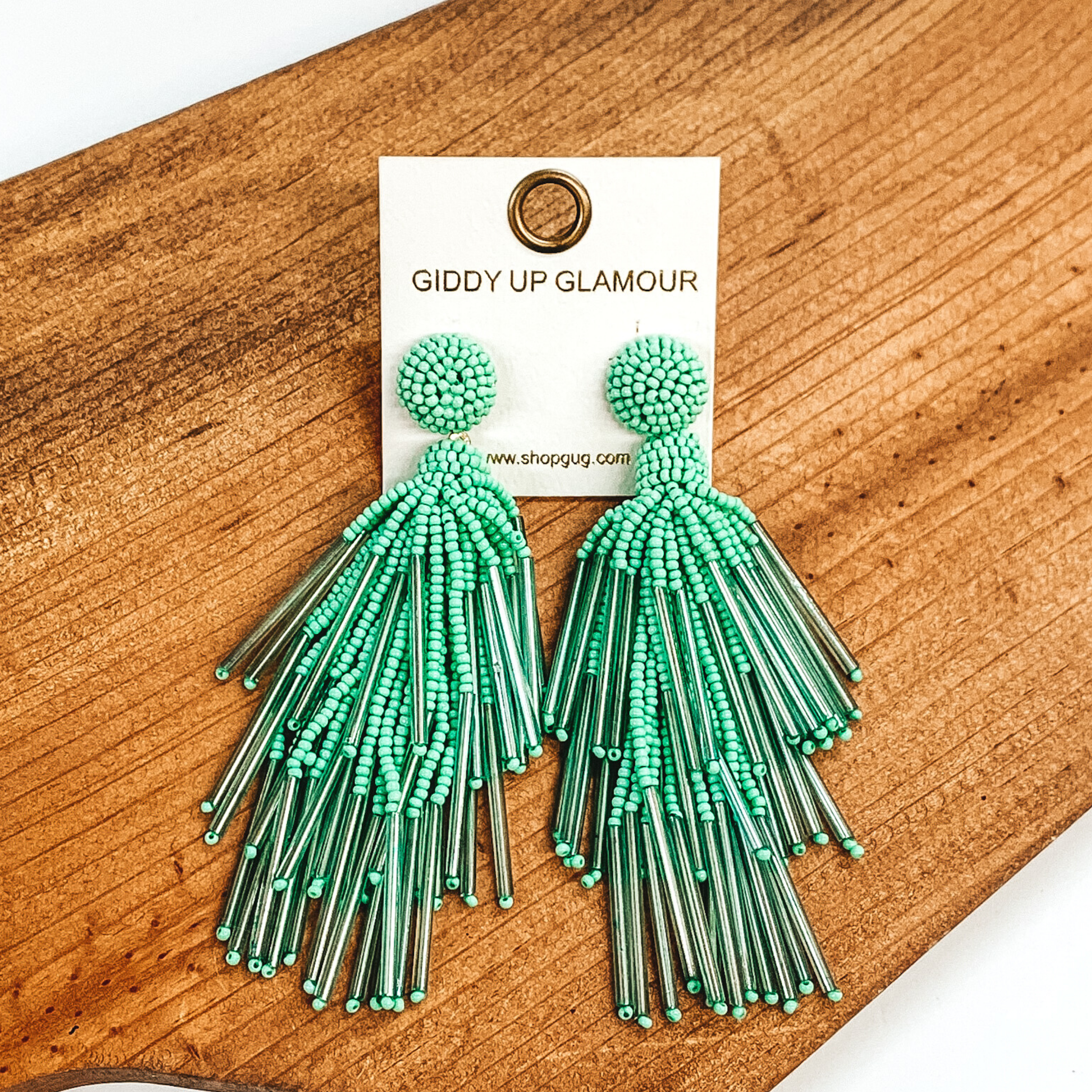 Circle beaded stud earrings with a beaded tassel in mint. These earrings are pictured laying on a brown piece of wood on a white background.