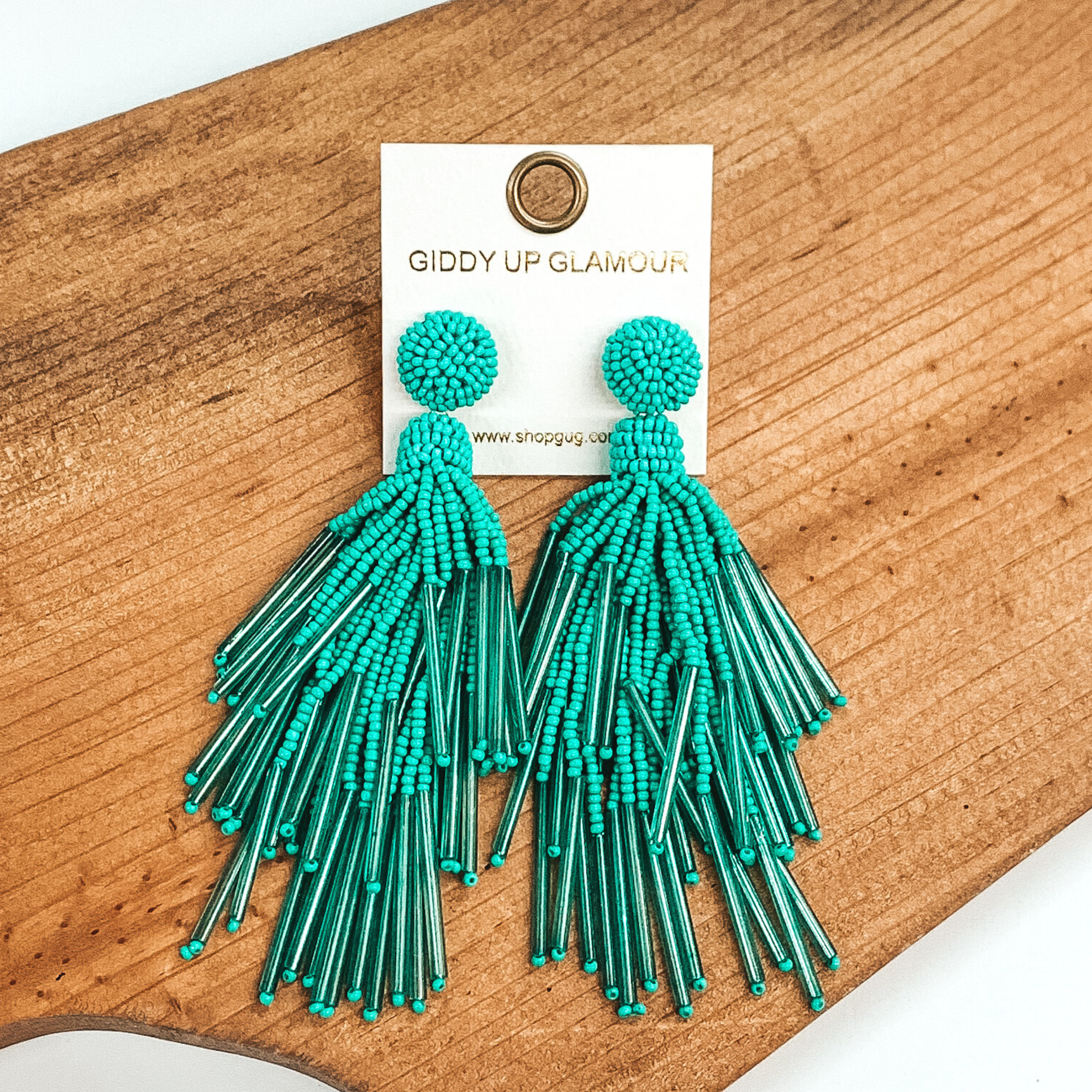 Circle beaded stud earrings with a beaded tassel in turquoise. These earrings are pictured laying on a brown piece of wood on a white background.