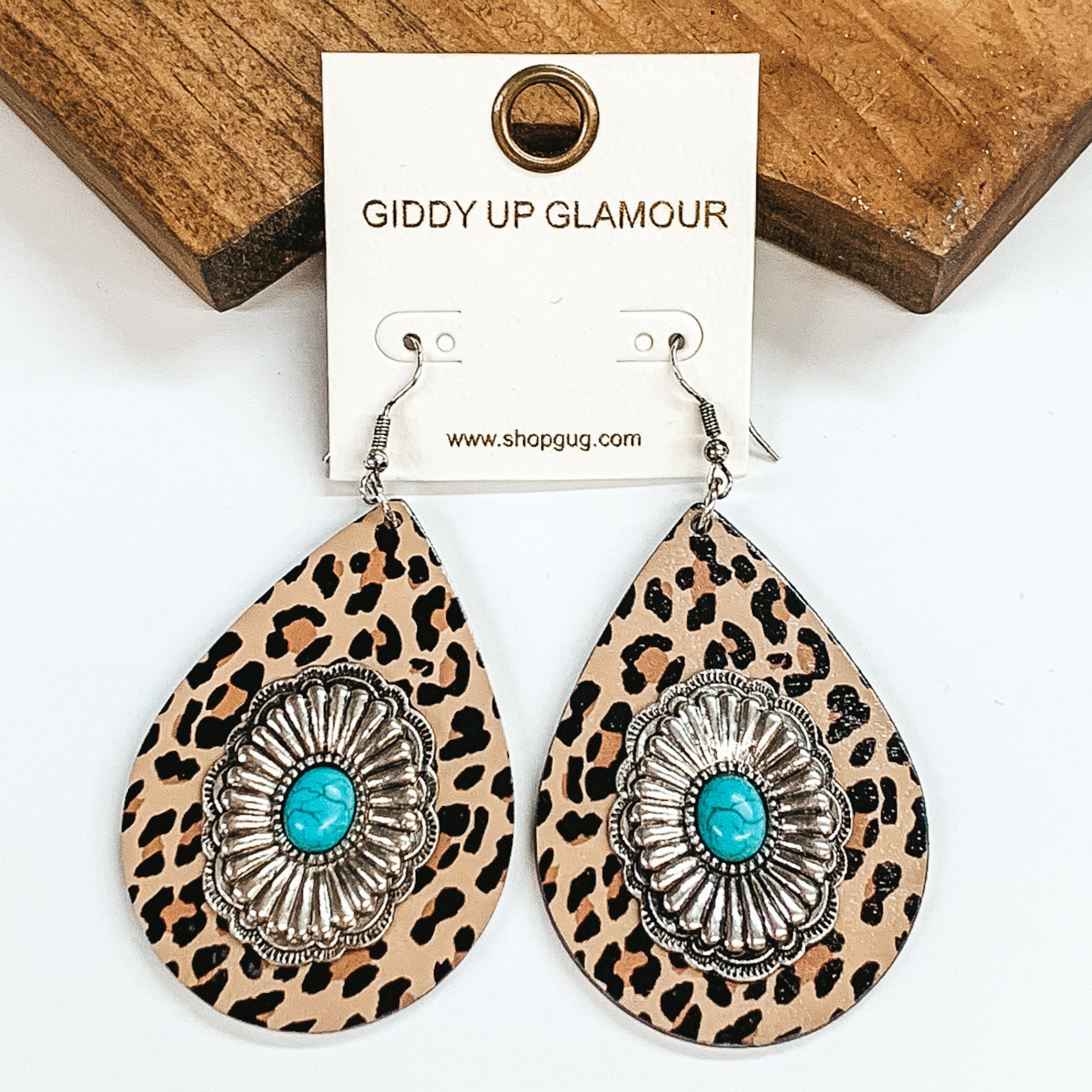 Teardrop earrings in brown cheetah print with silver conchos and turquoise stones in the center. Pictured on a white background and brown block.