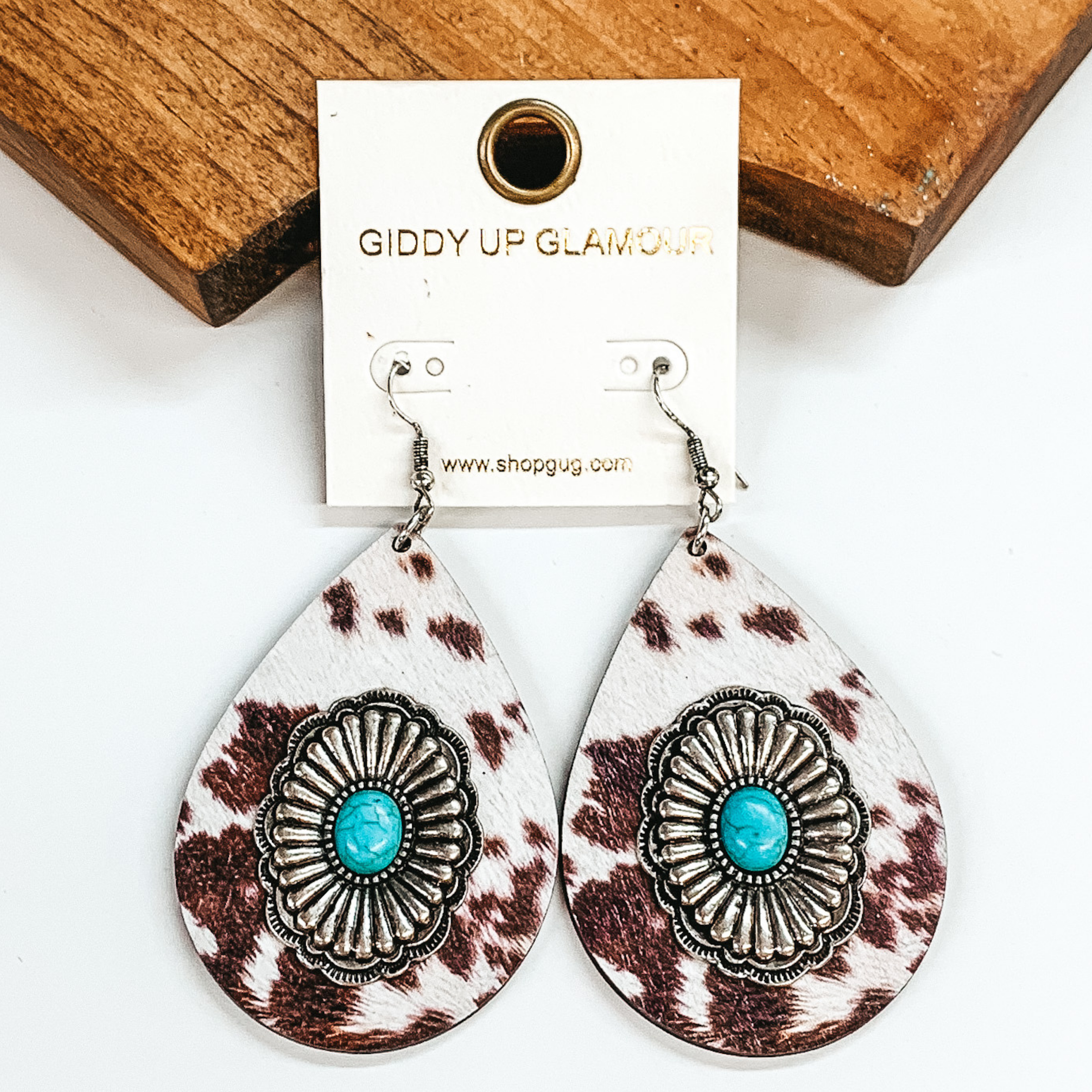 Teardrop earrings in brown cow print with silver conchos and turquoise stones in the center. Pictured on a white background and a brown block.