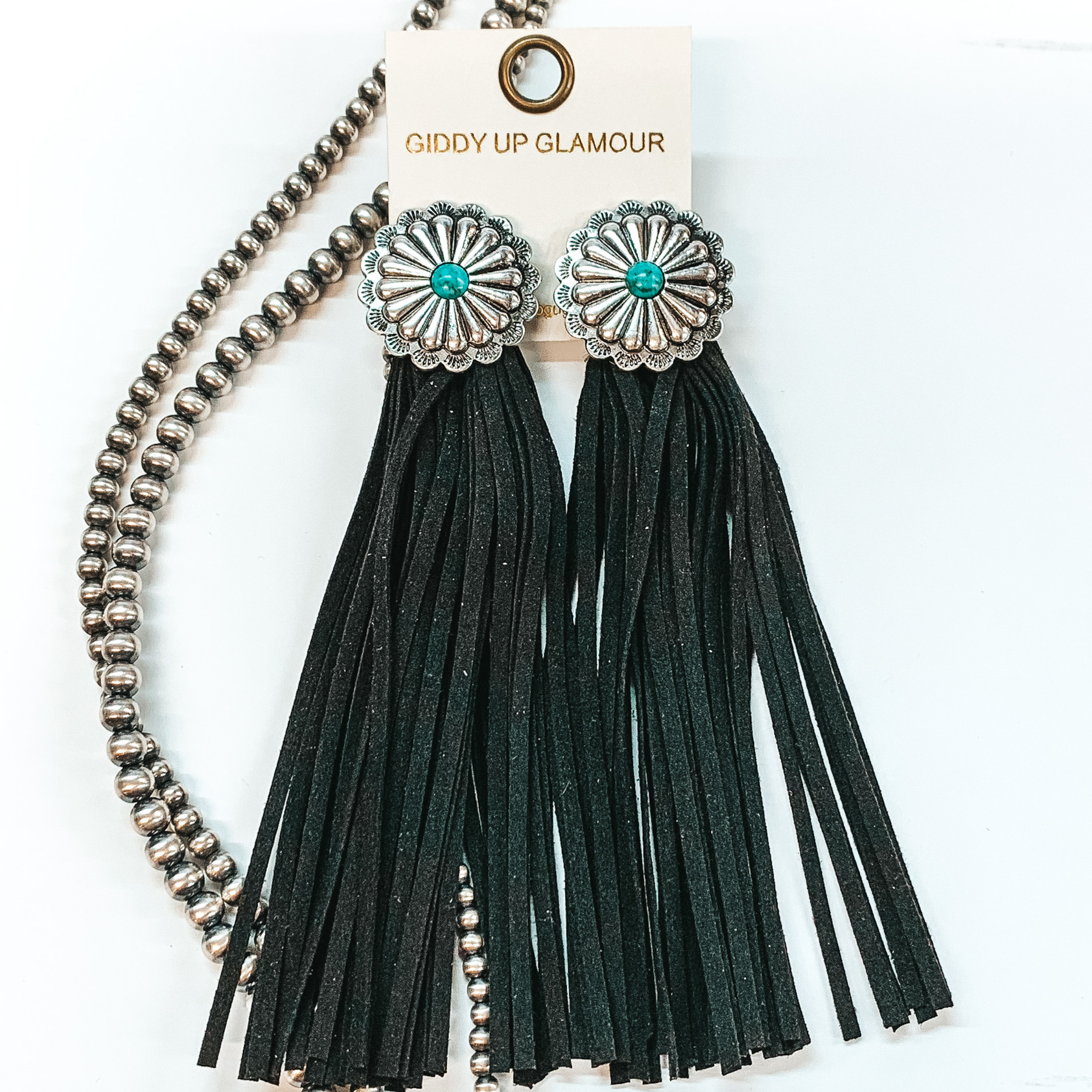 Post back concho earrings with a turquoise stone  in the center with black faux leather tassels. Pictured in a white background with beads as decor.