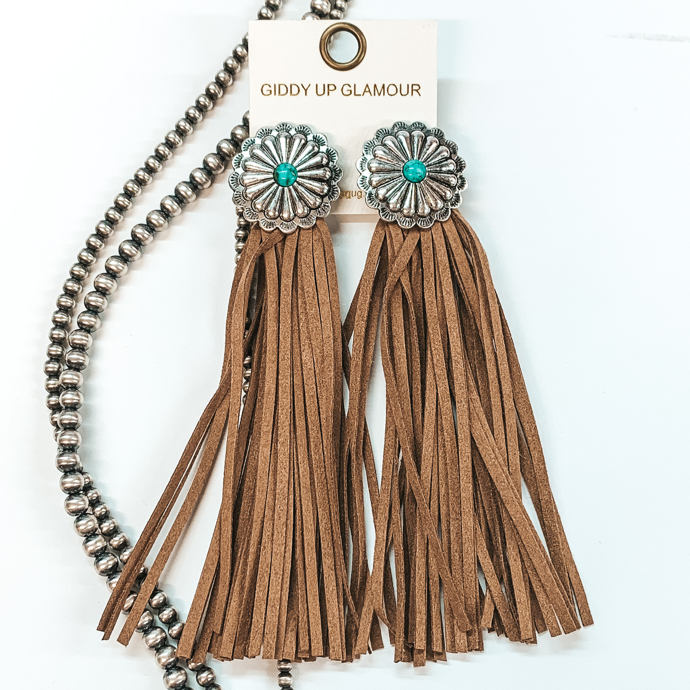 Post back concho earrings with a turquoise stone  in the center with brown faux leather tassels. Pictured in a white background with beads as decor.