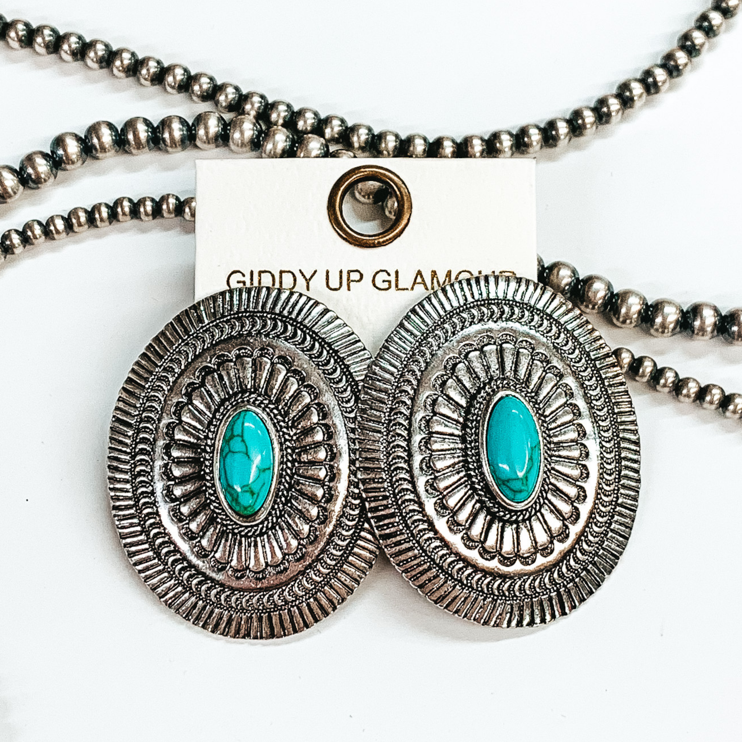 Large silver concho earrings with small oval turquoise stones in the center. Pictured in a white background with beads as decor.