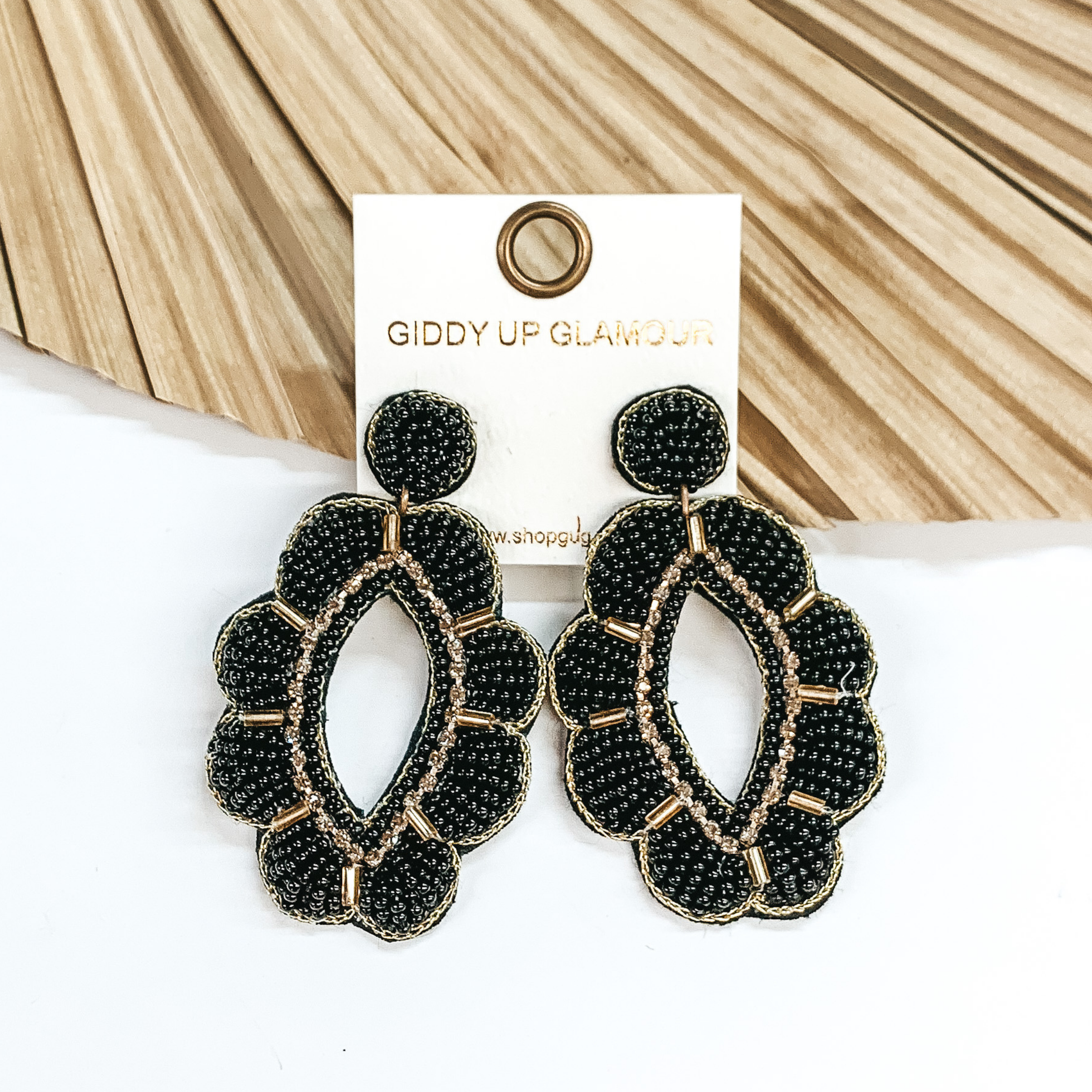 Black beaded earrings in a floral teardrop shape  with gold outline. Long gold beads in between  each petal with gold, shiny beads around in the middle. Taken in a white background with a light brown decor piece.