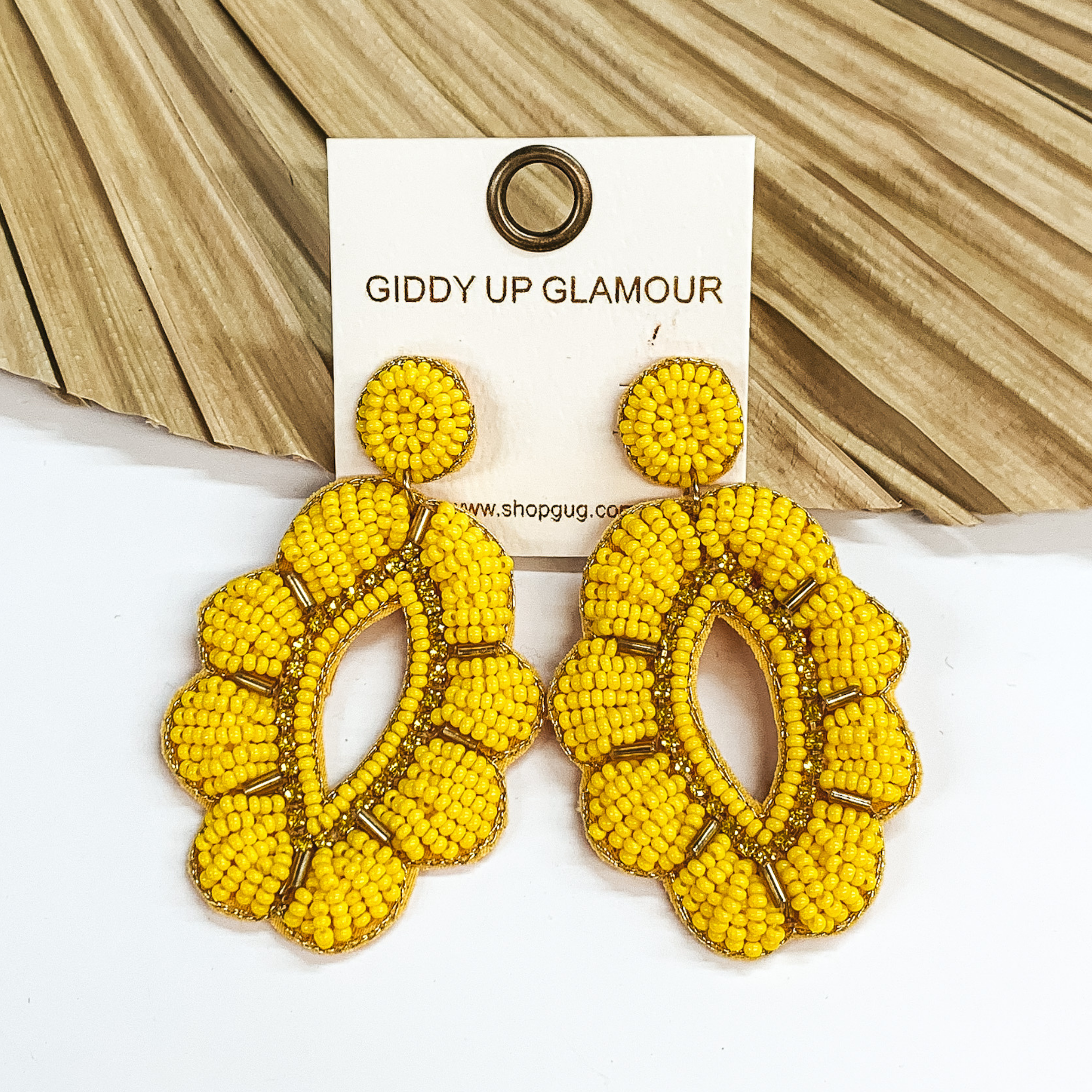 Yellow beaded earrings in a floral teardrop shape  with gold outline. Long gold beads in between  each petal with yellow, shiny beads around in  the middle.  Taken in a white background with a light brown decor piece.