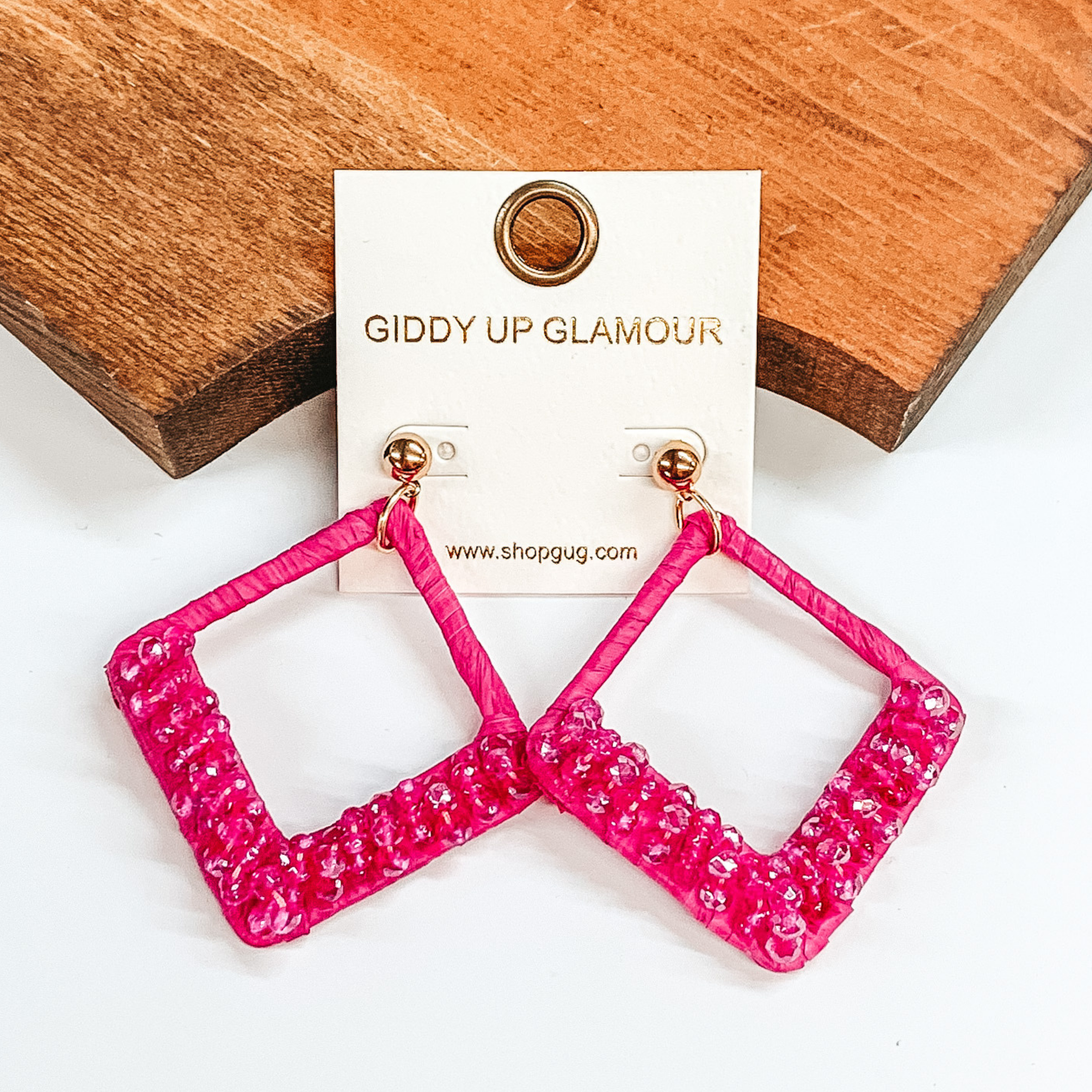 Open diamond shaped dangle earrings wrapped in  raffia that includes a layer of beads on the  bottom half of the earrings. These earrings are  pink. These earrings are pictured on on a  white background and a brown block.