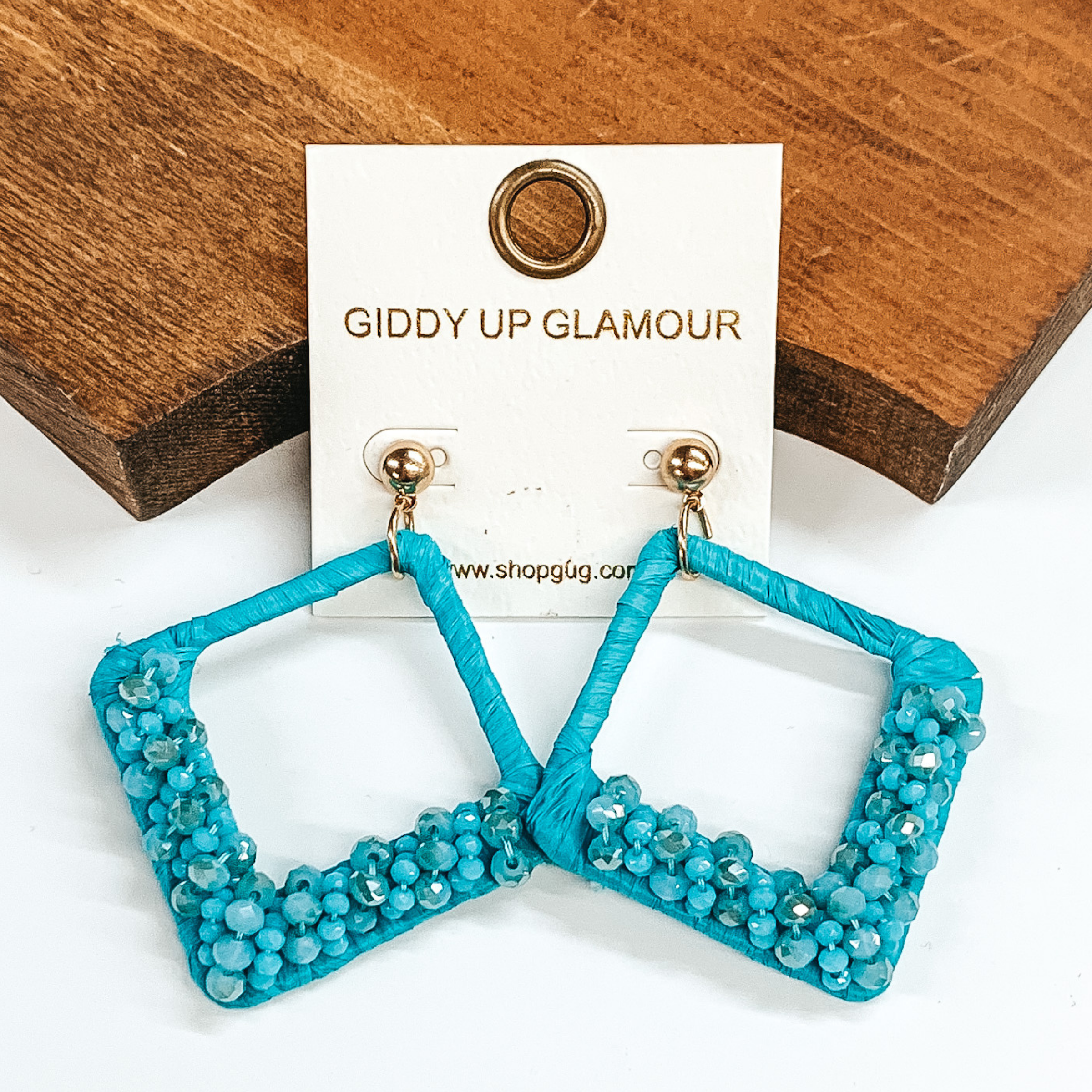 Open diamond shaped dangle earrings wrapped in  raffia that includes a layer of beads on the  bottom half of the earrings. These earrings are  blue. These earrings are pictured on on a  white background and a brown block.