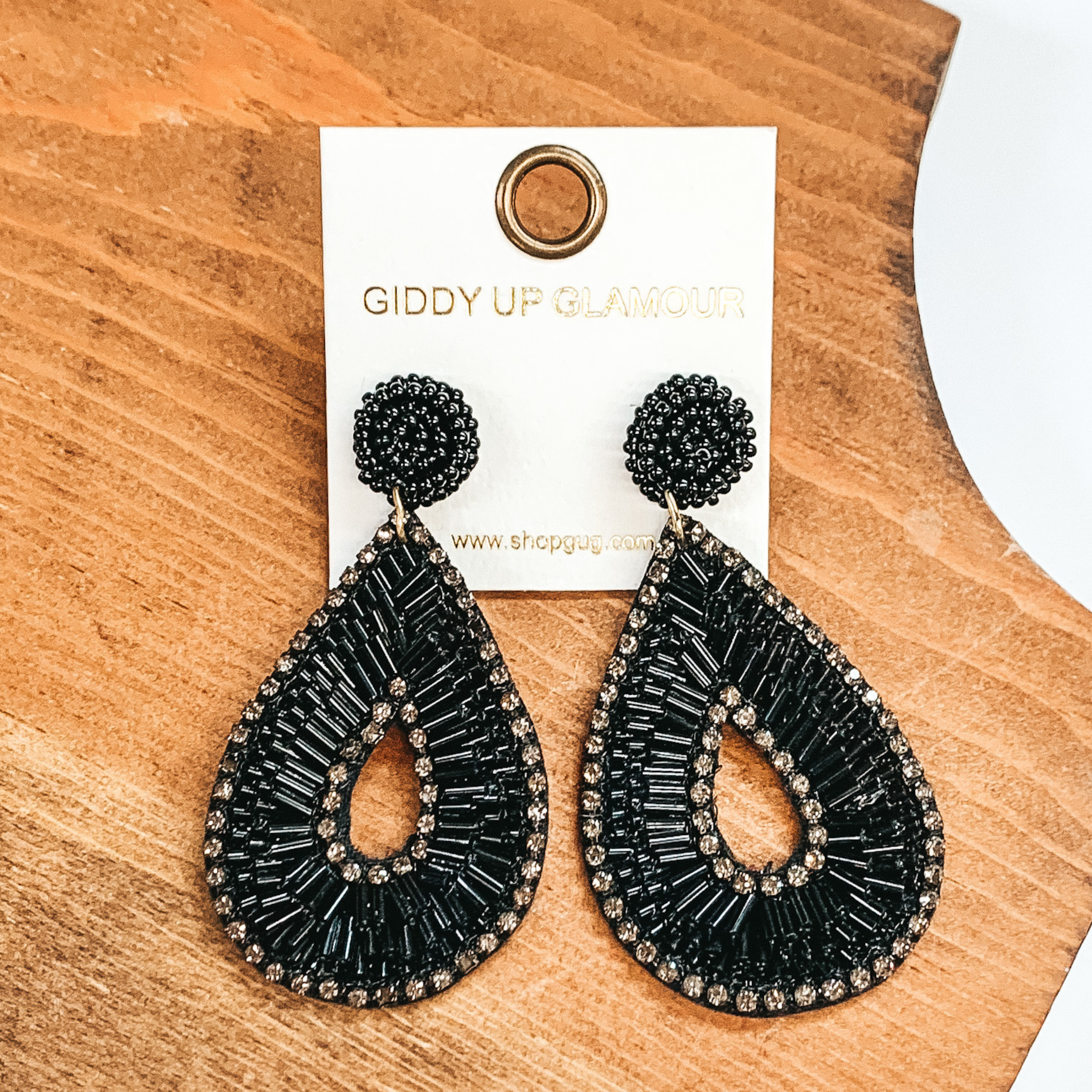 Black bugle beaded earrings in teardrop shape. Outlined with dark grey crystals and in the middle, with black bugle beads in between. These earrings  are pictured on a brown block and a white background.