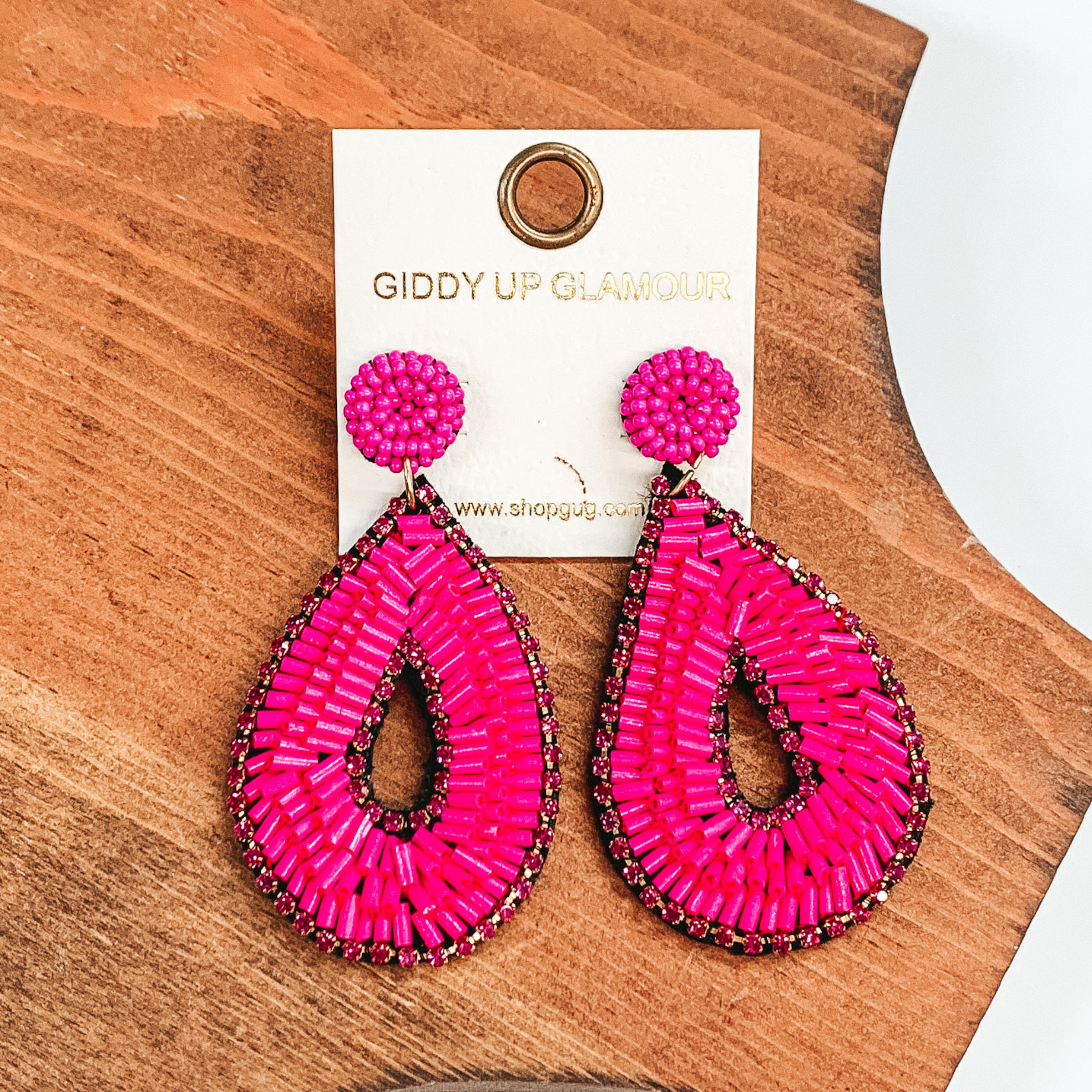 Pink bugle beaded earrings in teardrop shape. Outlined with pink crystals and in the middle, with pink bugle beads in between. These earrings  are pictured on a brown block and a white background.