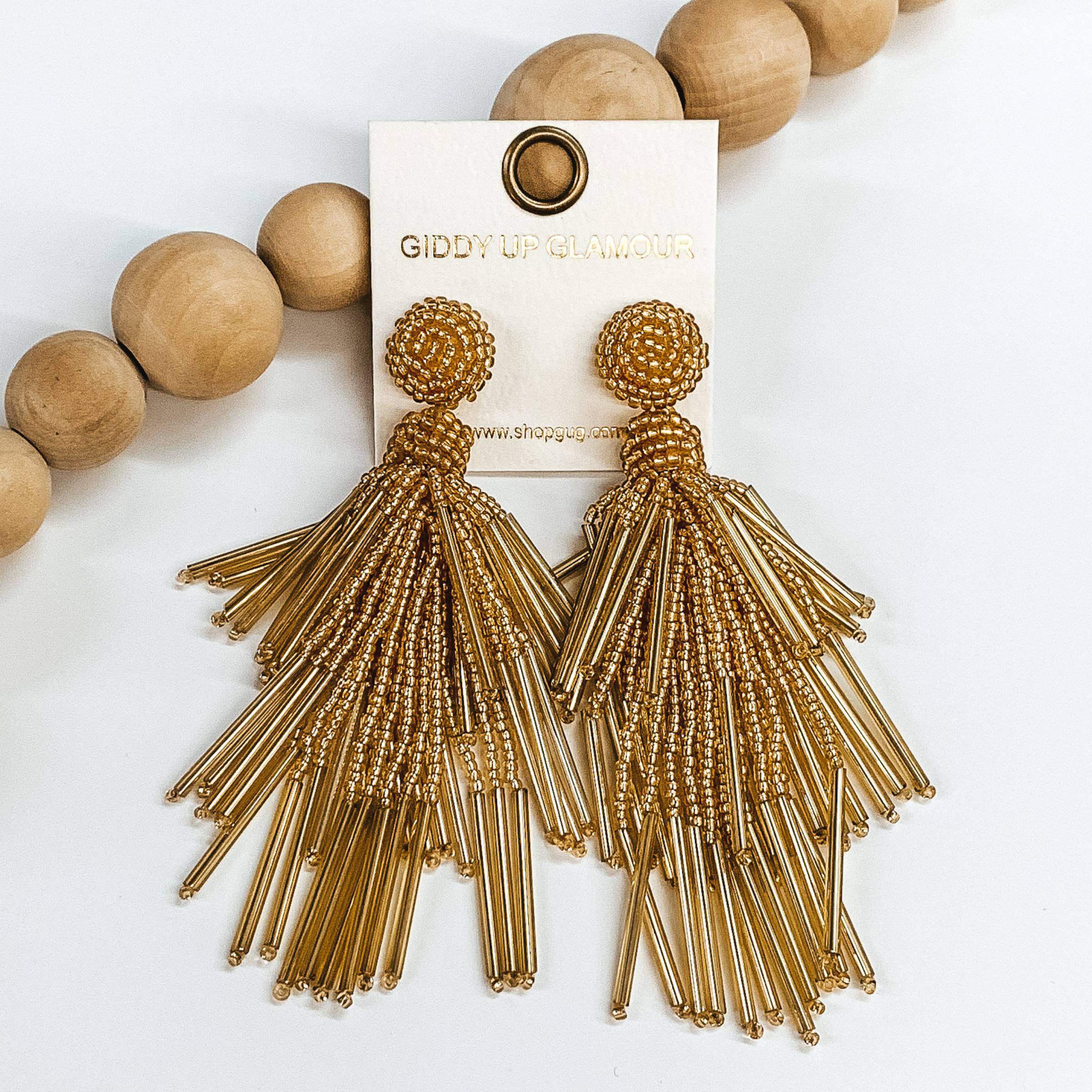 Circle beaded stud earrings with a beaded  tassel in gold. These earrings are pictured  laying on a white background with brown beads as  decor.