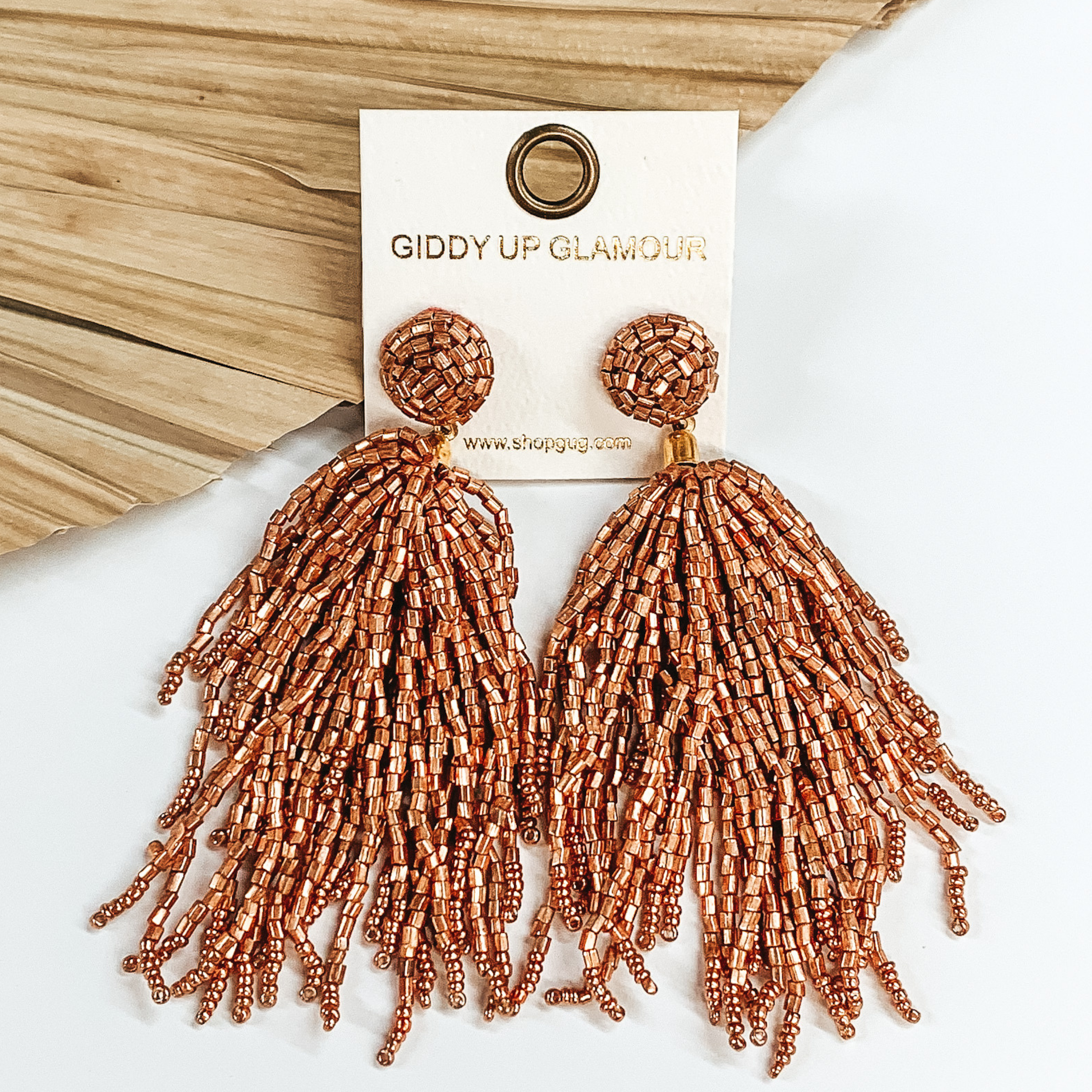 Long beaded stud earrings with a beaded  tassel in rose gold. These earrings are pictured  on a white background with a dried palm leaf  in the background.