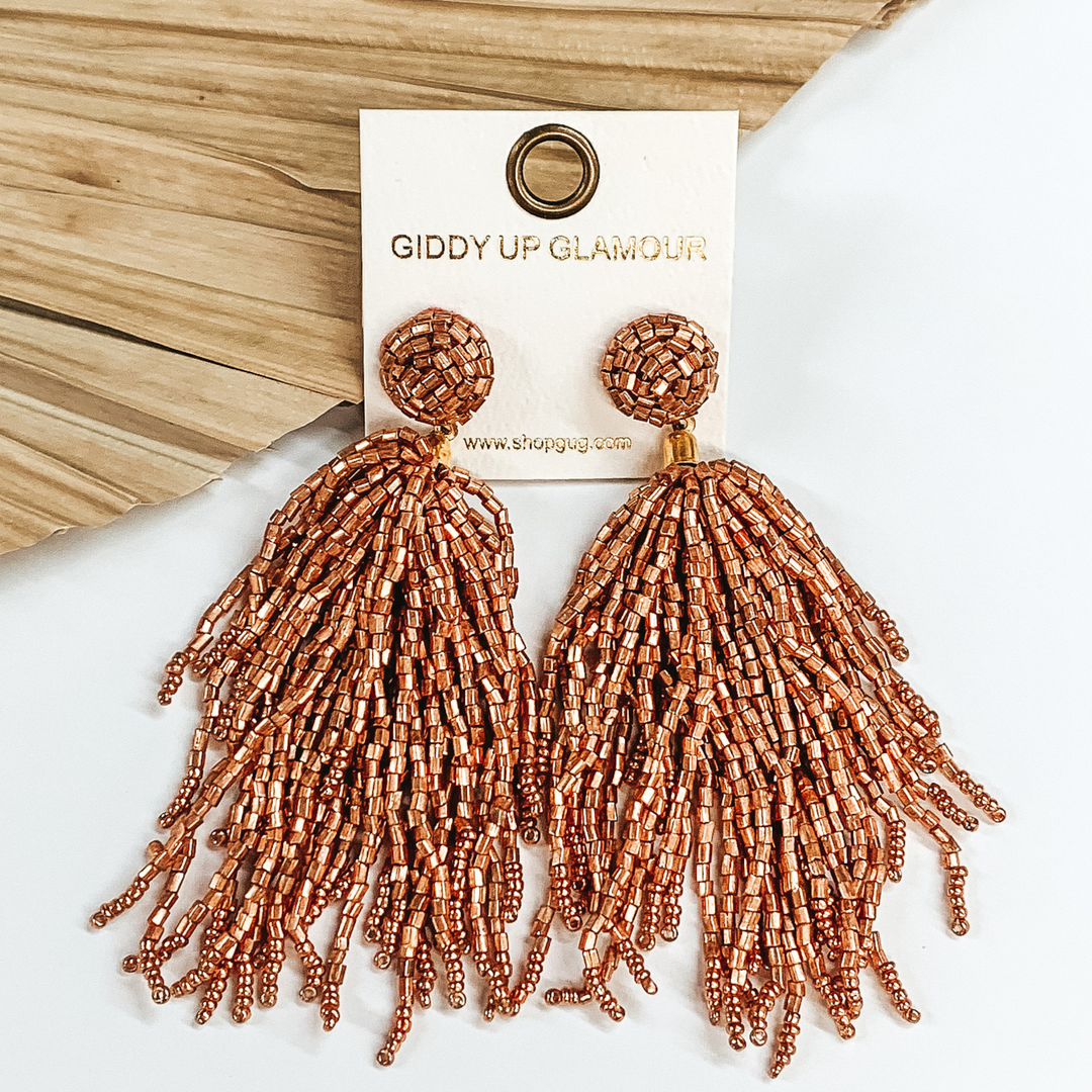 Long beaded stud earrings with a beaded  tassel in rose gold. These earrings are pictured  on a white background with a dried palm leaf  in the background.