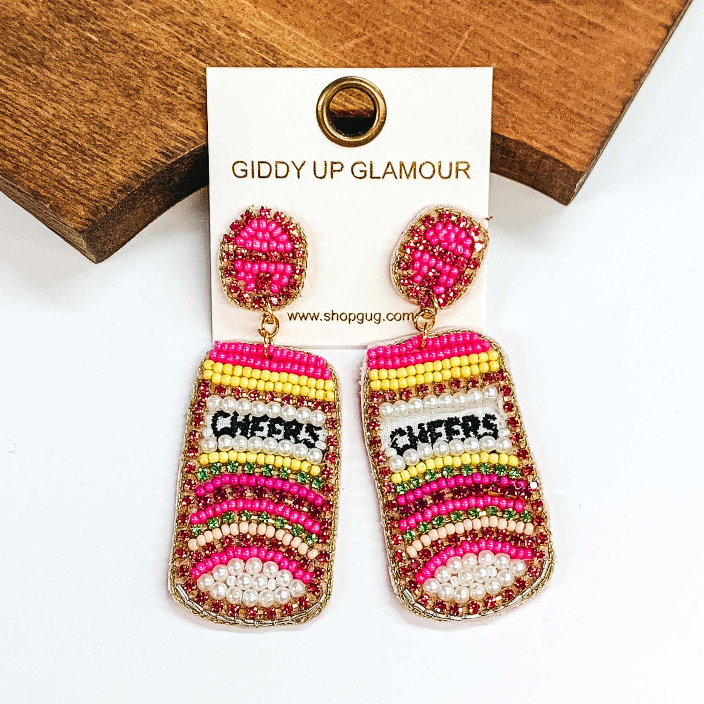 Drinking can beaded earrings in different shades of  pink beads and yellow. With green,light pink, and hot pink crystals, and pearls. With "CHEERS" in the  center. Outlined with gold thread. Taken in a white  background with a brown block.