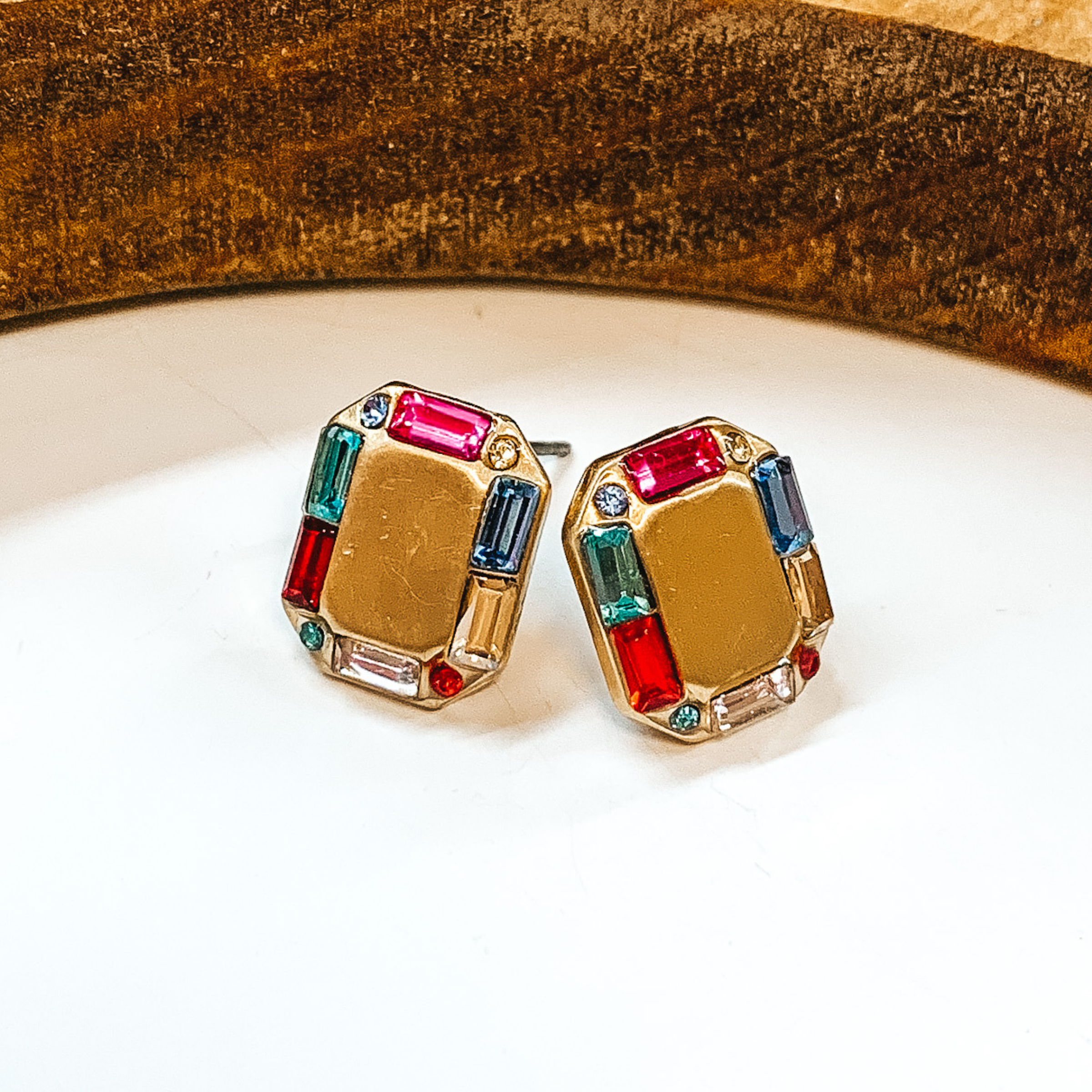 Gold rectangle shaped earrings with a crystal  multicolor outline. Small multicolor crystals in  pink, teal, blue, red. light pink, and clear  outlining it.  Taken on a white background with a brown block  in the back.