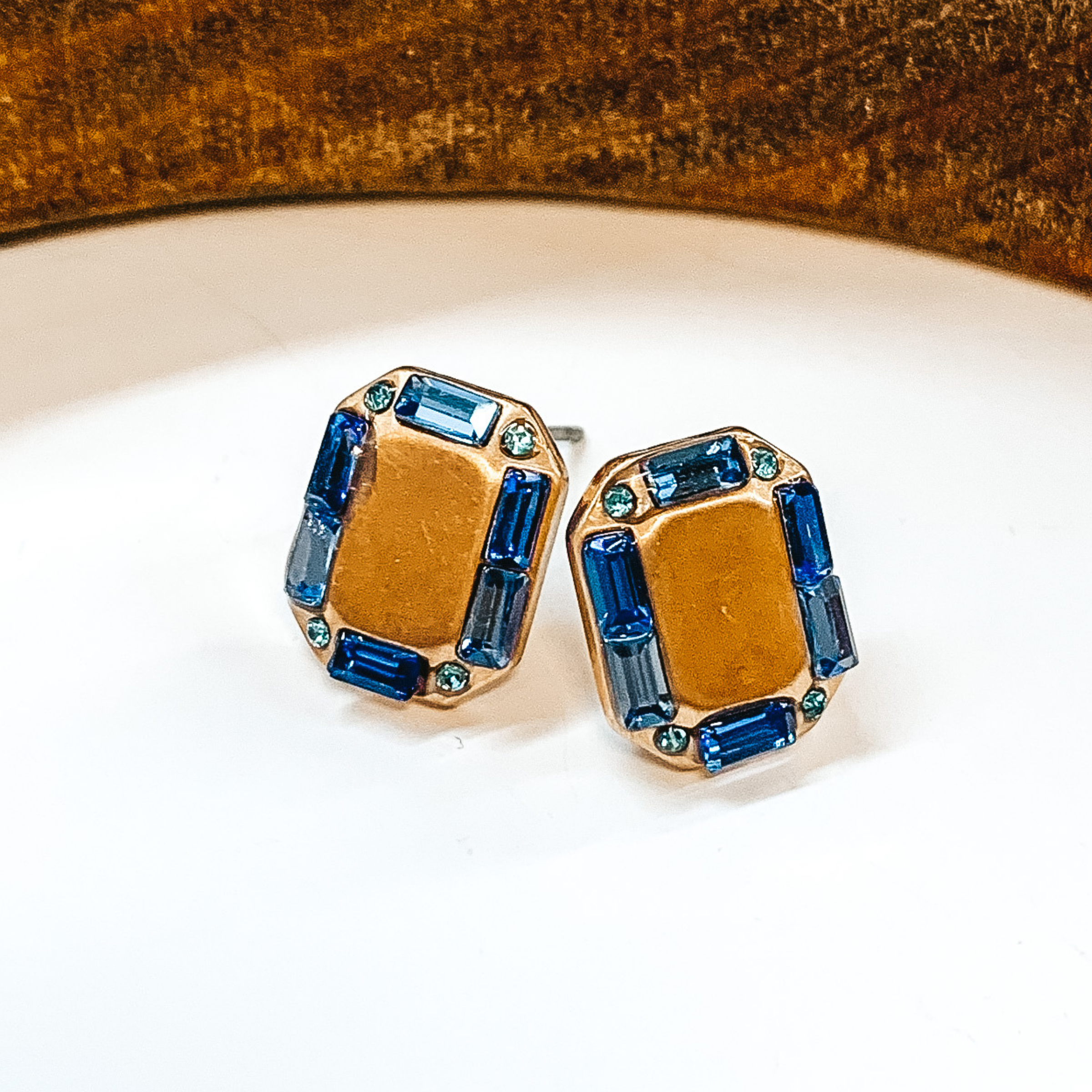 Gold rectangle shaped earrings with a blue  crystal outline. Small blue crystals in different  tones. Taken on a white background with a brown  block in the back.