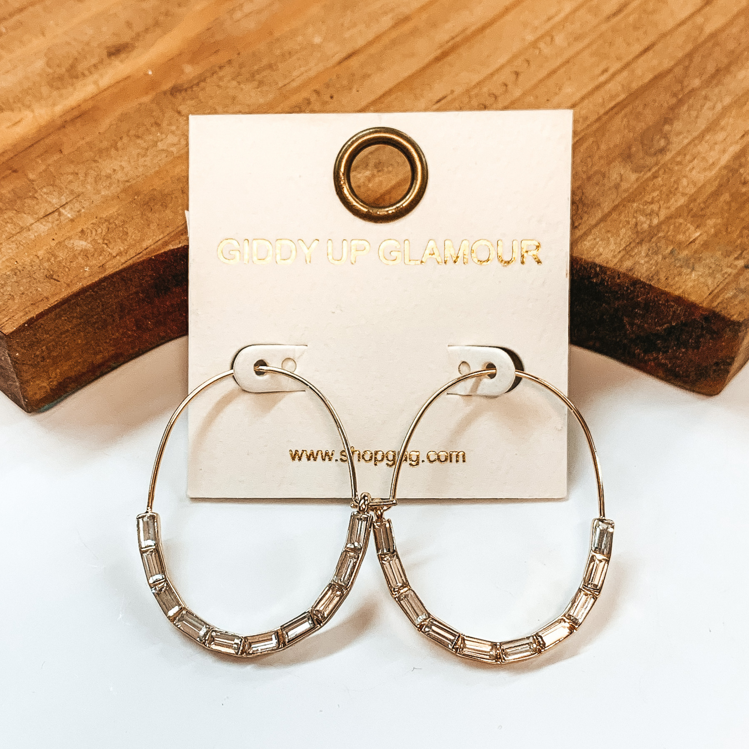 Gold oval hoops with clear, long crystals in  the bottom half. Taken on a white background with a  brown block in the back.