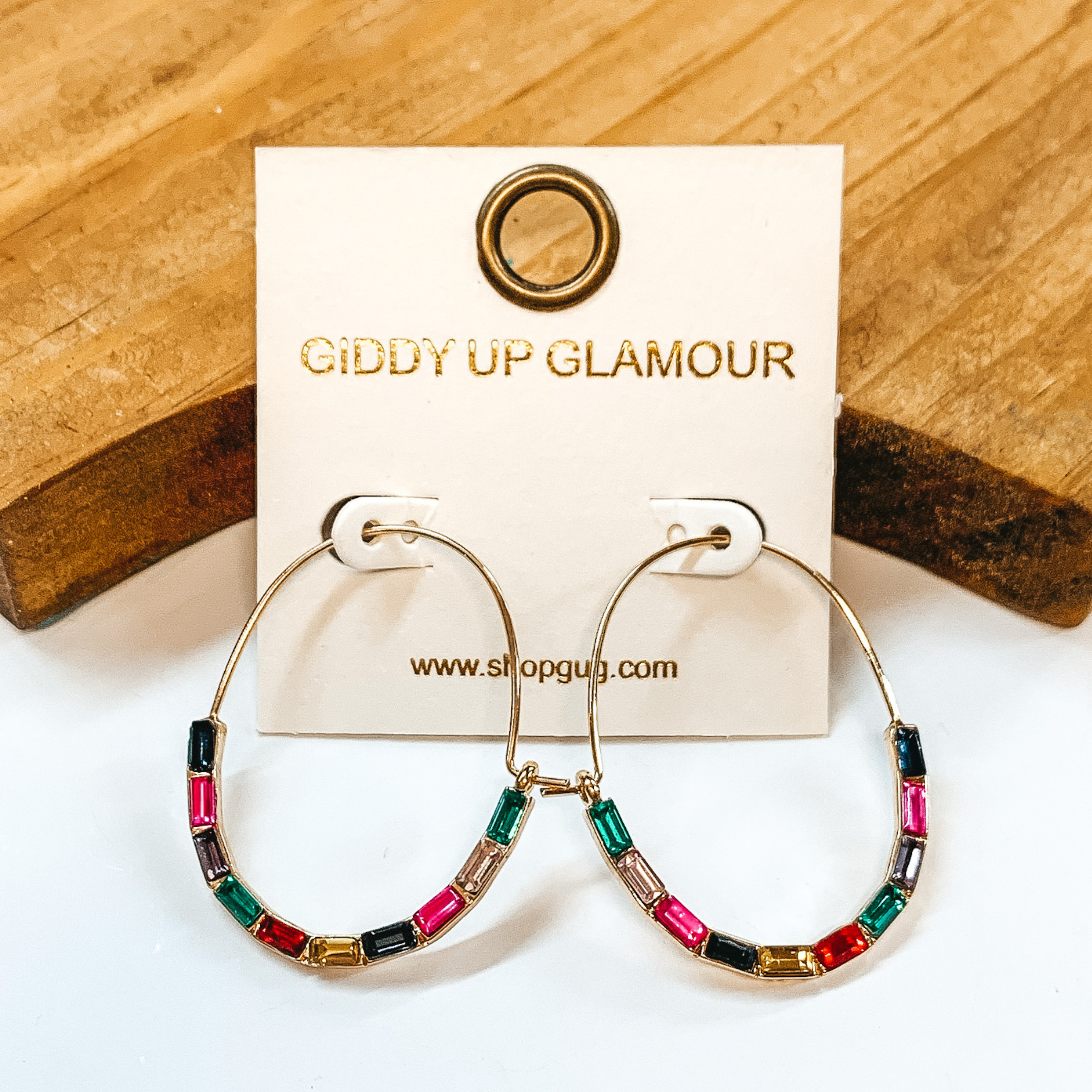 Gold oval hoops with multicolor, long crystals in  the bottom half. Multicolor crystals in  dark blue, hot pink, purple, green, red, and yellow. Taken on a white background with a  brown block in the back.