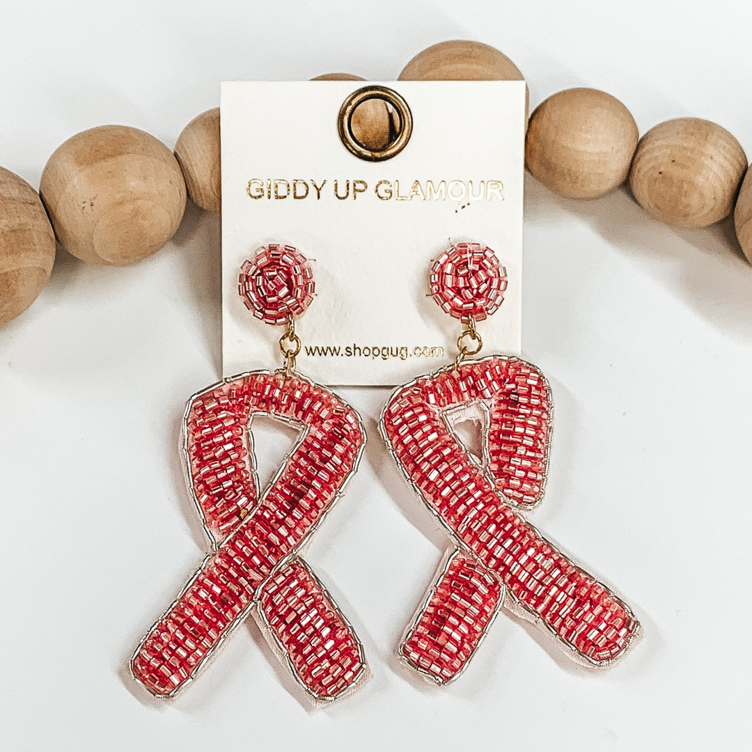 Champagne pink beaded circle studs with hanging  beaded ribbon pendant and silver thread  lining. These earrings are pictured  on a white background with brown beads in the back  as decor.