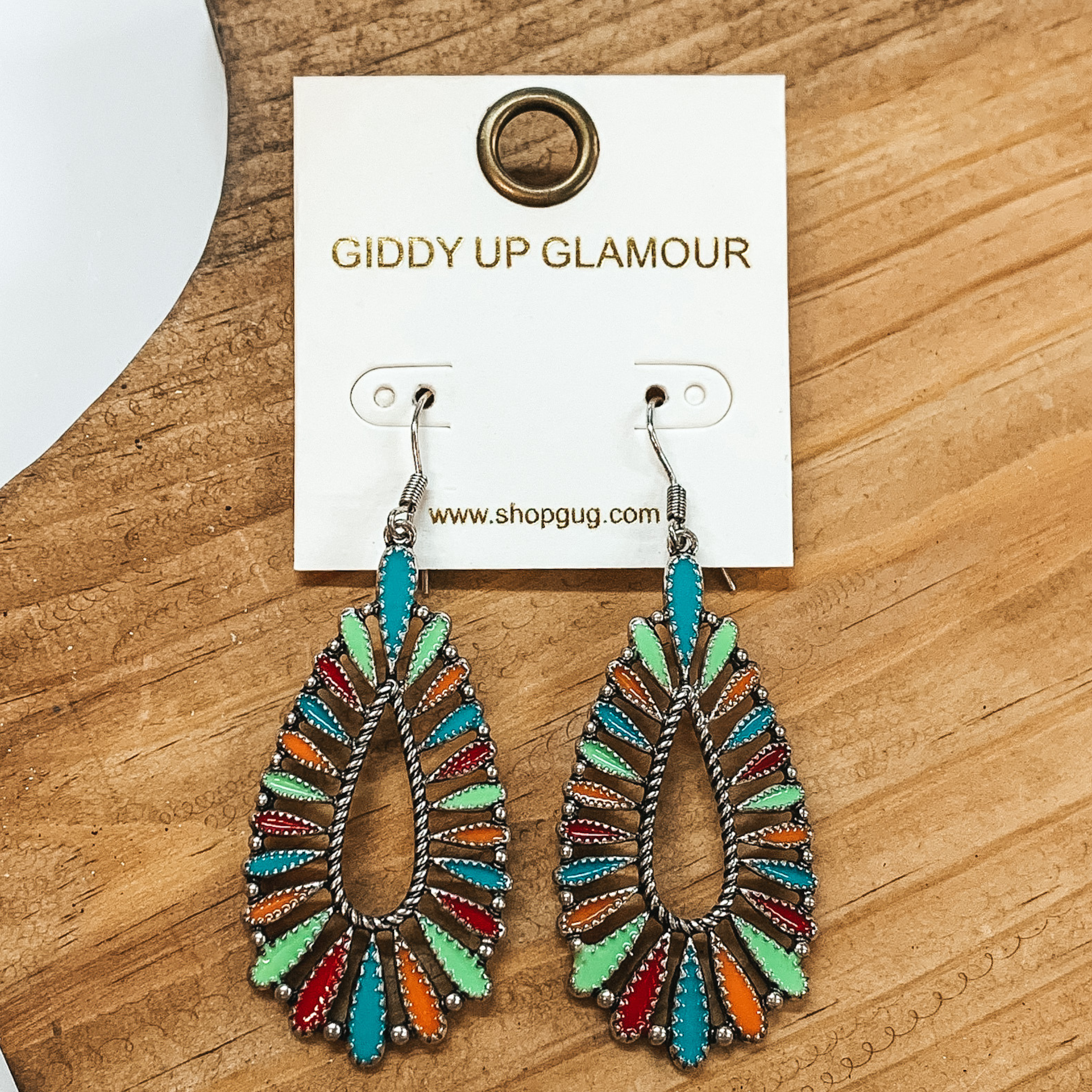 Teardrop earrings in multicolored needle points.  Colors in turquoise, green, orange, and red with a  silver outline. These earrings are pictured on a  brown block and white background.