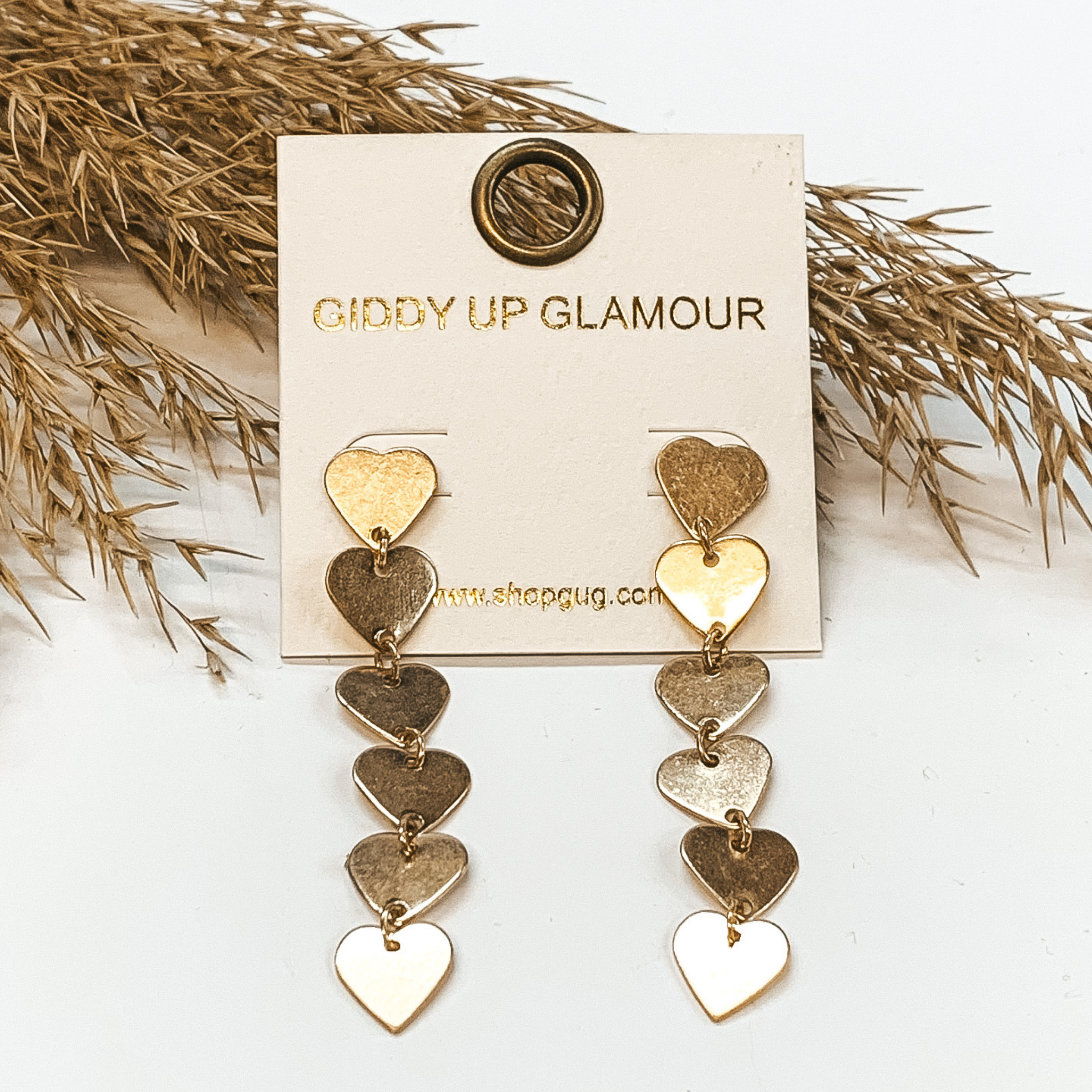 Drop earrings with six gold hearts on each earring. These earrings are pictured on a white background with a a brown plant in the back as decor.