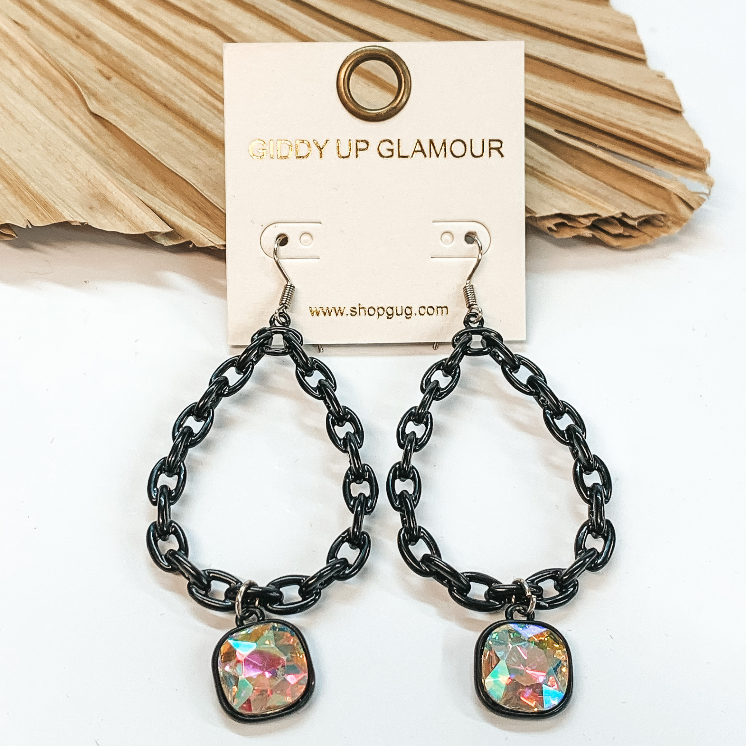 Black, chained, teardrop dangle earrings with a cushion cut drop AB crystal. These earrings are pictured on a white background with a brown leaf  in the back as decor.