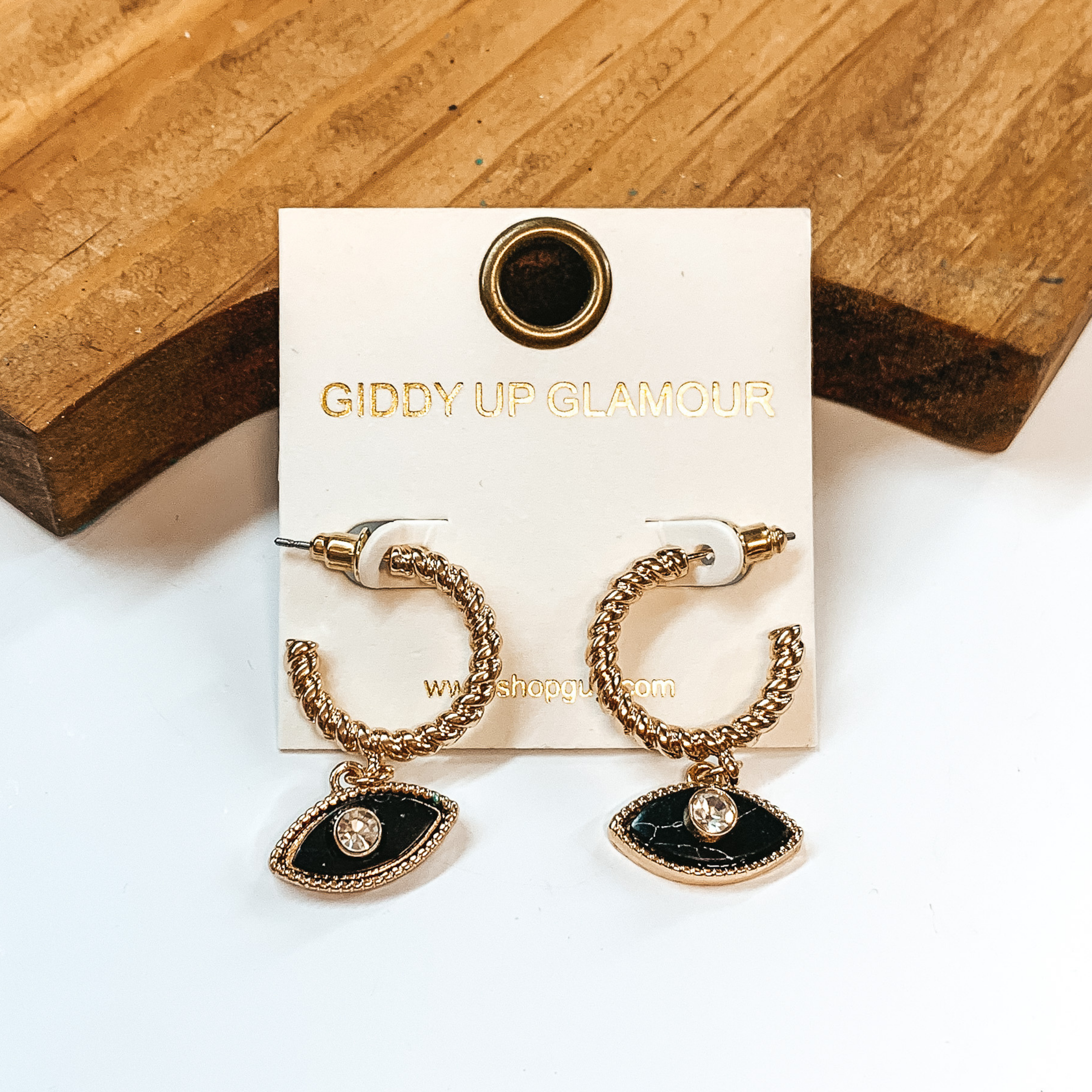 Small gold hoop earrings with a rope texture all  around. With a small evil eye pendant in black marble with a cz crystal in the middle, outlined in gold. Taken on a white background and a brown block in  the back.