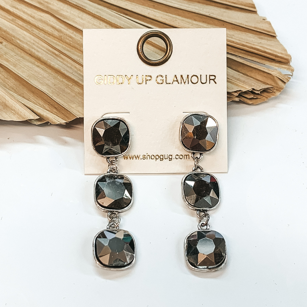 Silver drop earrings with three silver  cushion cut crystals. These earrings are pictured on a white background with a dried  palm leaf in the background.