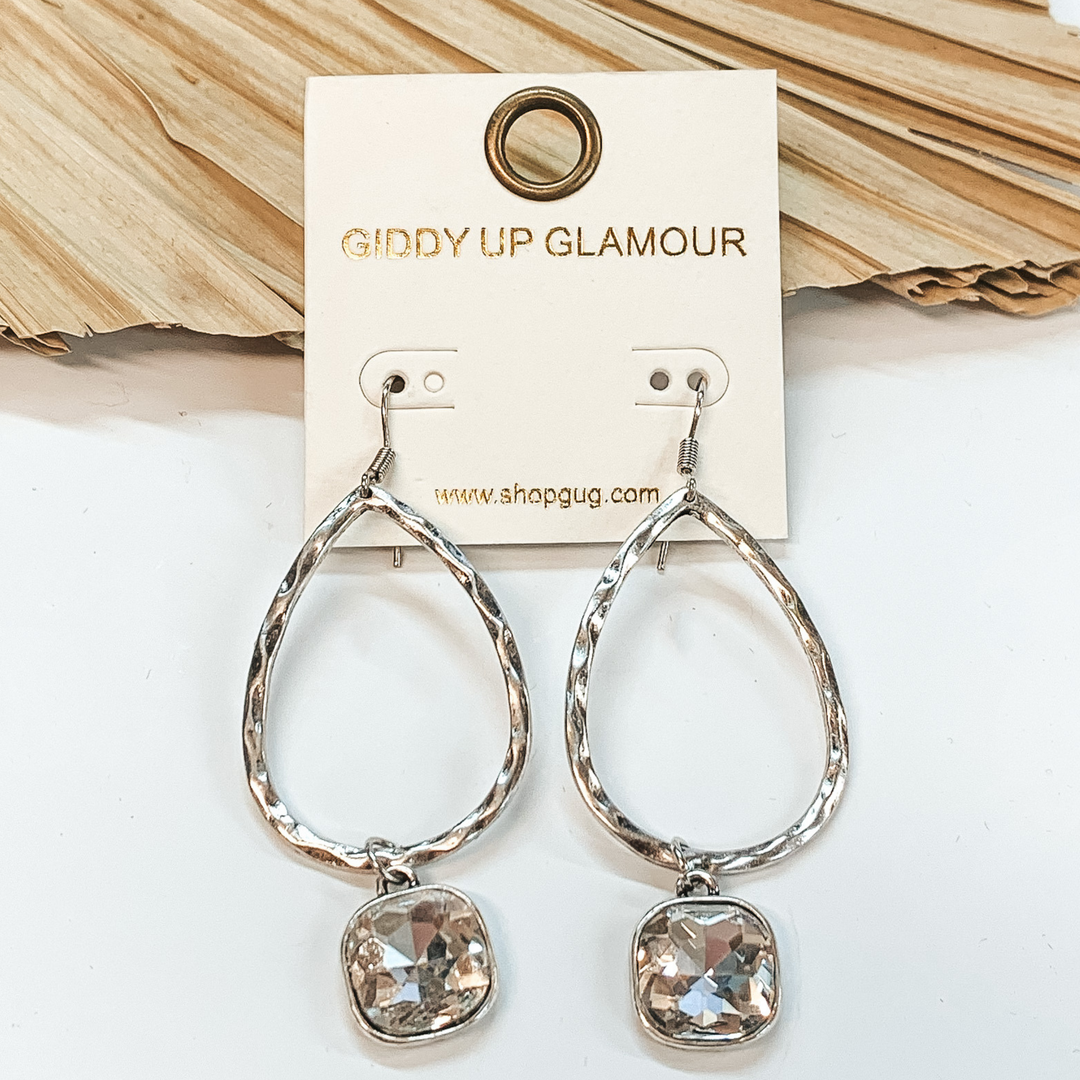 Silver teardrop hammered earrings with a hanging  cushion cut clear crystal. These earrings are pictured on a white background with a dried  palm leaf in the background.