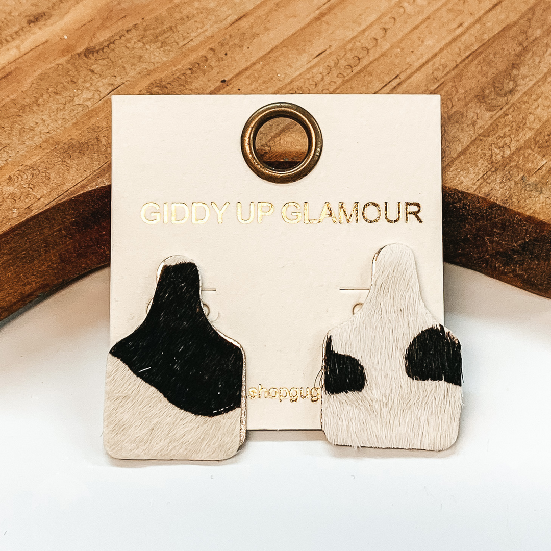 Cattle tag stud earrings with a hair on hide material.  It has a cow print in colors black and white. These earrings are pictured on a white background with a brown block in the back.