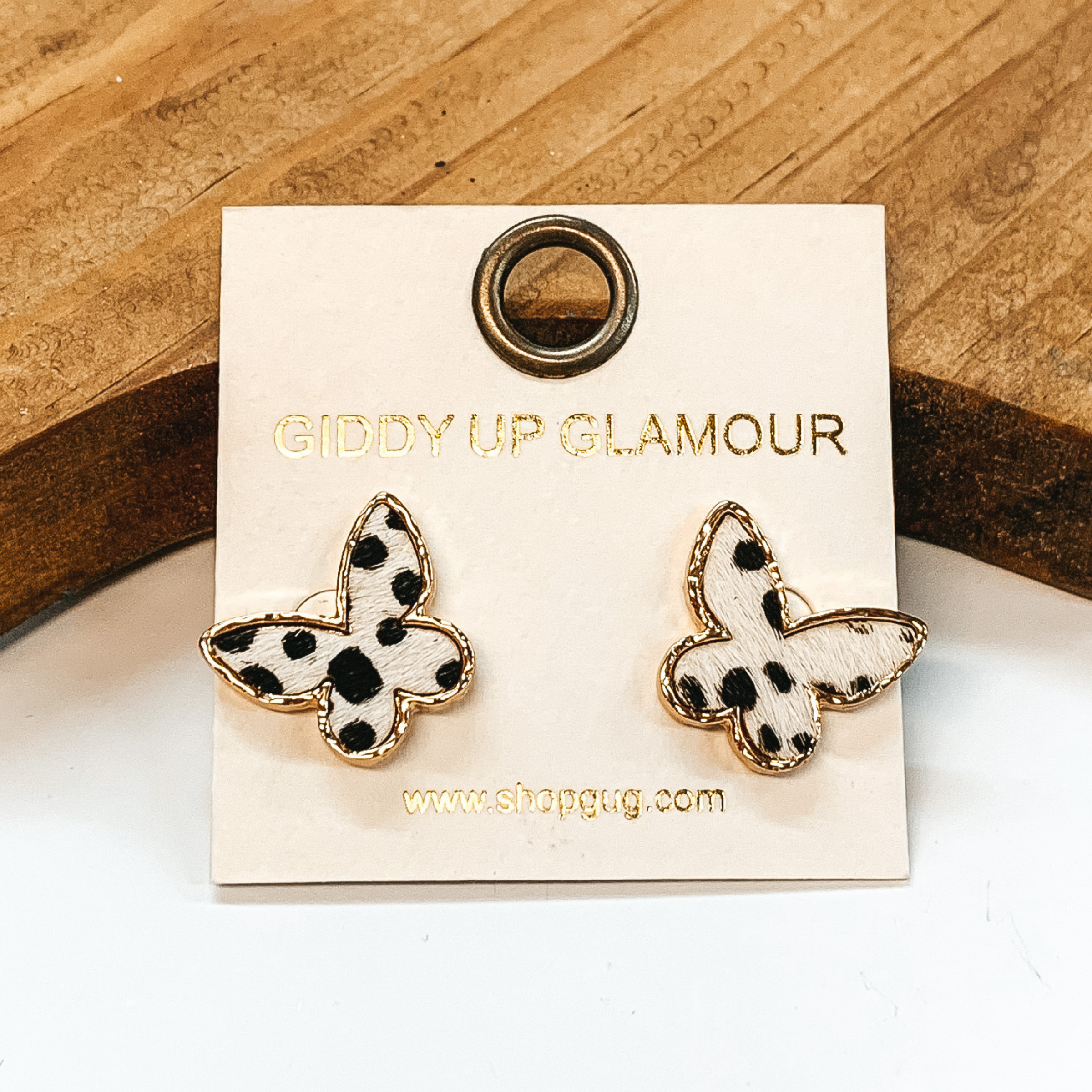 Gold butterfly studded earrings with genuine leather in white dotted print. These earrings are pictured on a white background with a brown block in the back.