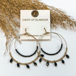 Double hoop earings in gold, inner hoop has small black beads. Outer hoop has small  gold beads with five black crystals. These earrings  are pictured on a white background with a brown  plant in the back as decor.