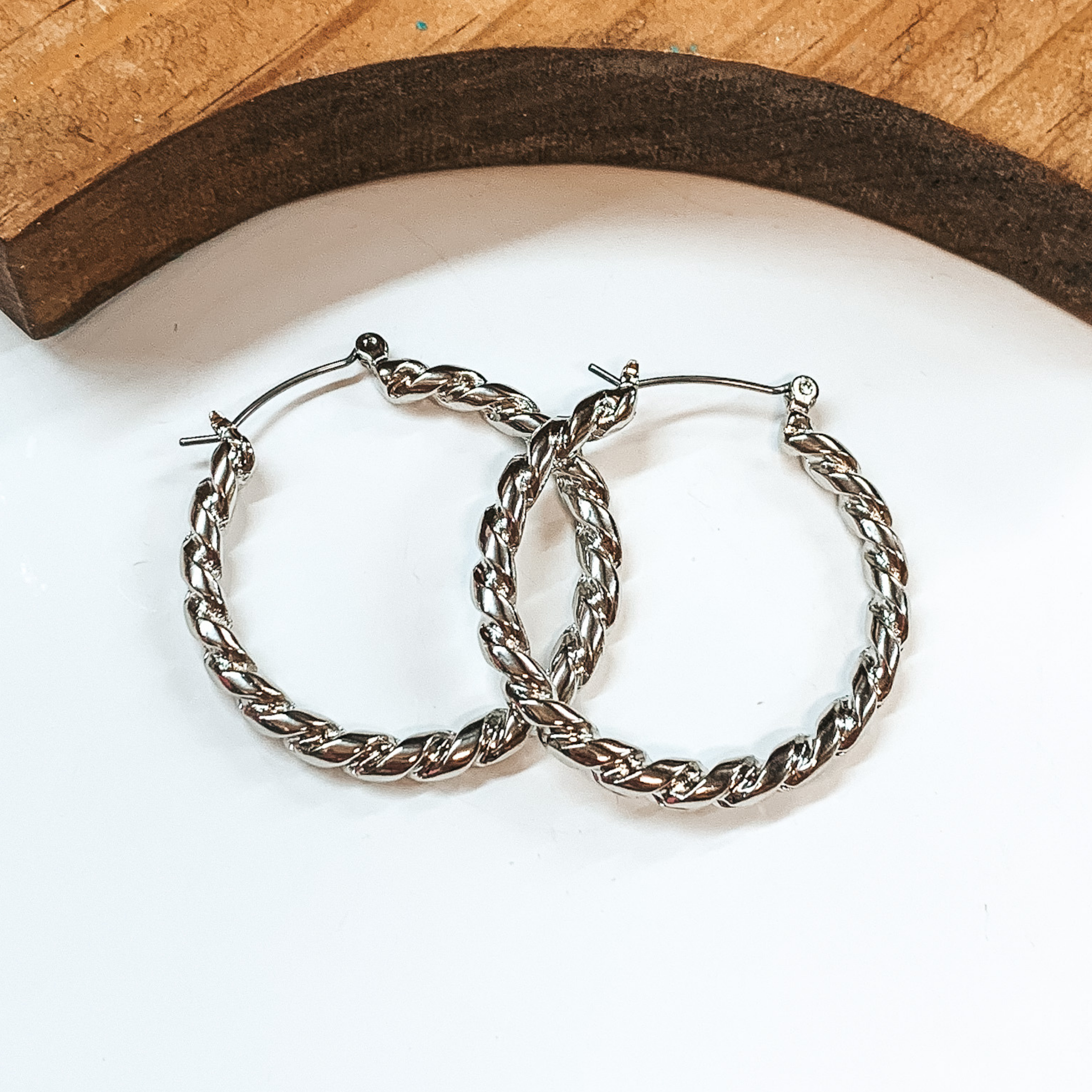 Silver hoop earrings with twisted rope texture. Taken on a white background with a brown block in the back.