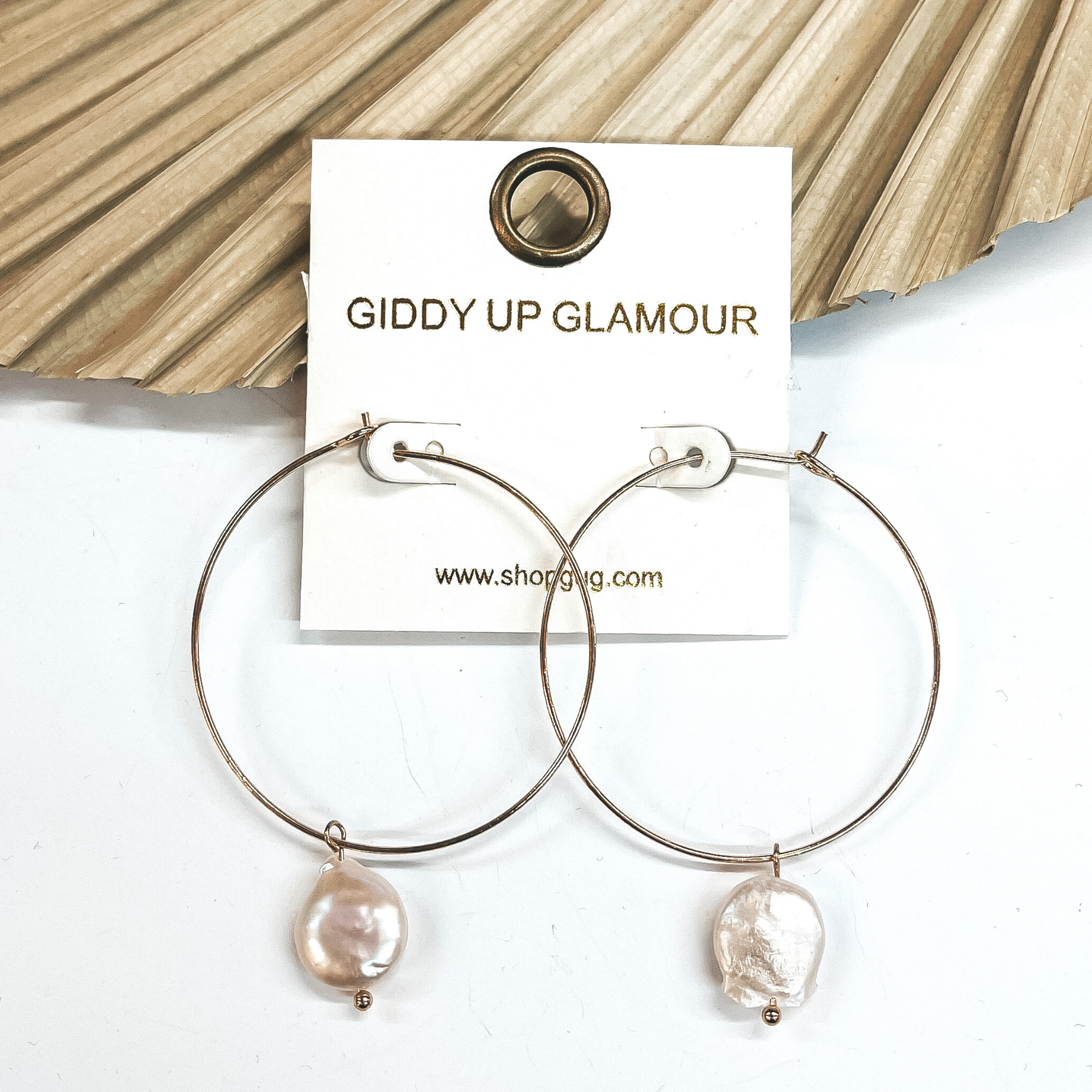 Thin gold wired hoop earings that includes a  pearl charm in the center. These earrings  are pictured on a white background with a  dried up palm leaf in the back as decor.