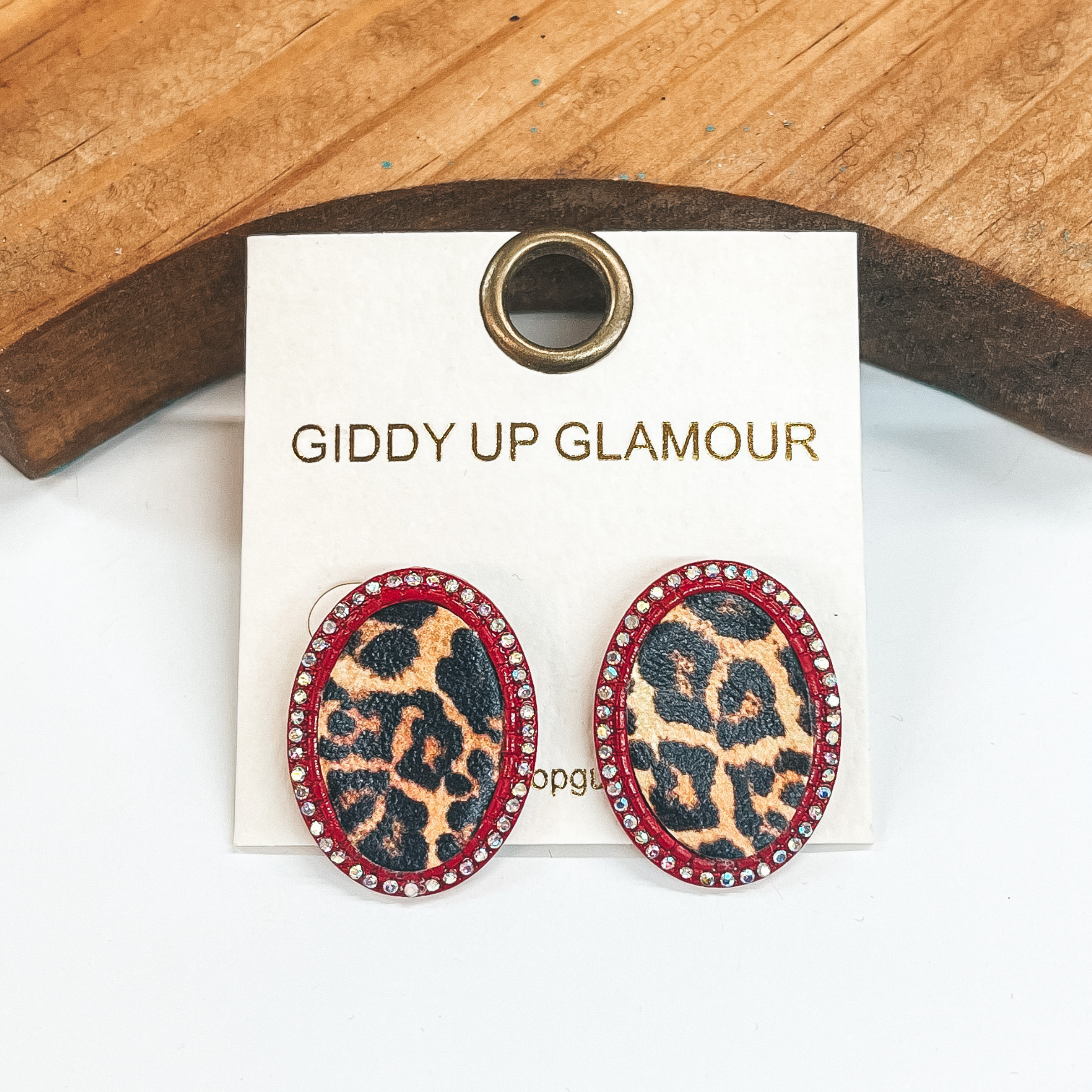 Red oval post back earrings with small  AB crystals all around. Leopard print in an oval  shape in the middle. These earrings  are pictured on a white background with a brown block in the back.