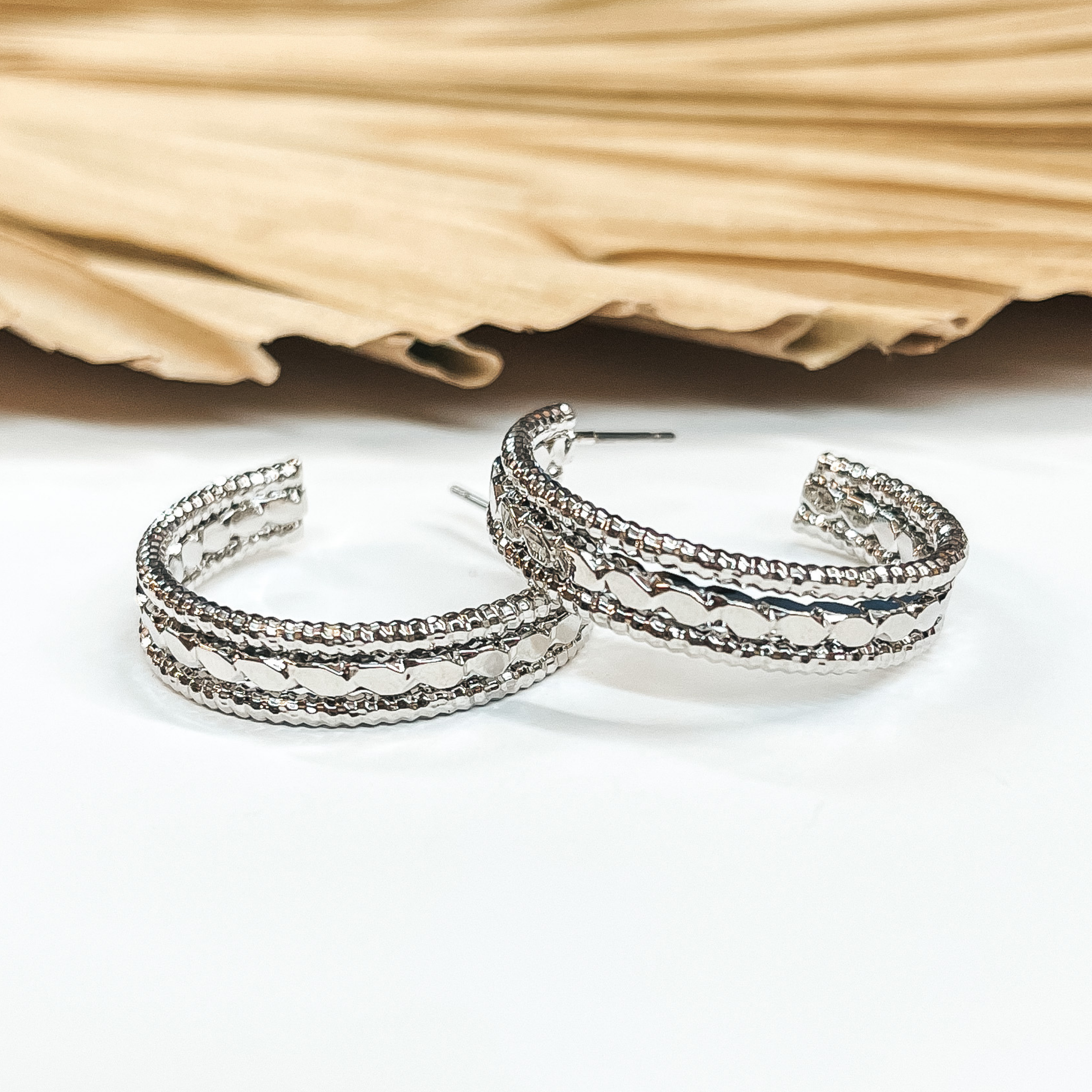 Open ended hoop earrings in silver with  different rope textures. These earrings are  pictured on a white background with a brown, dried up palm leaf in the back as decor.