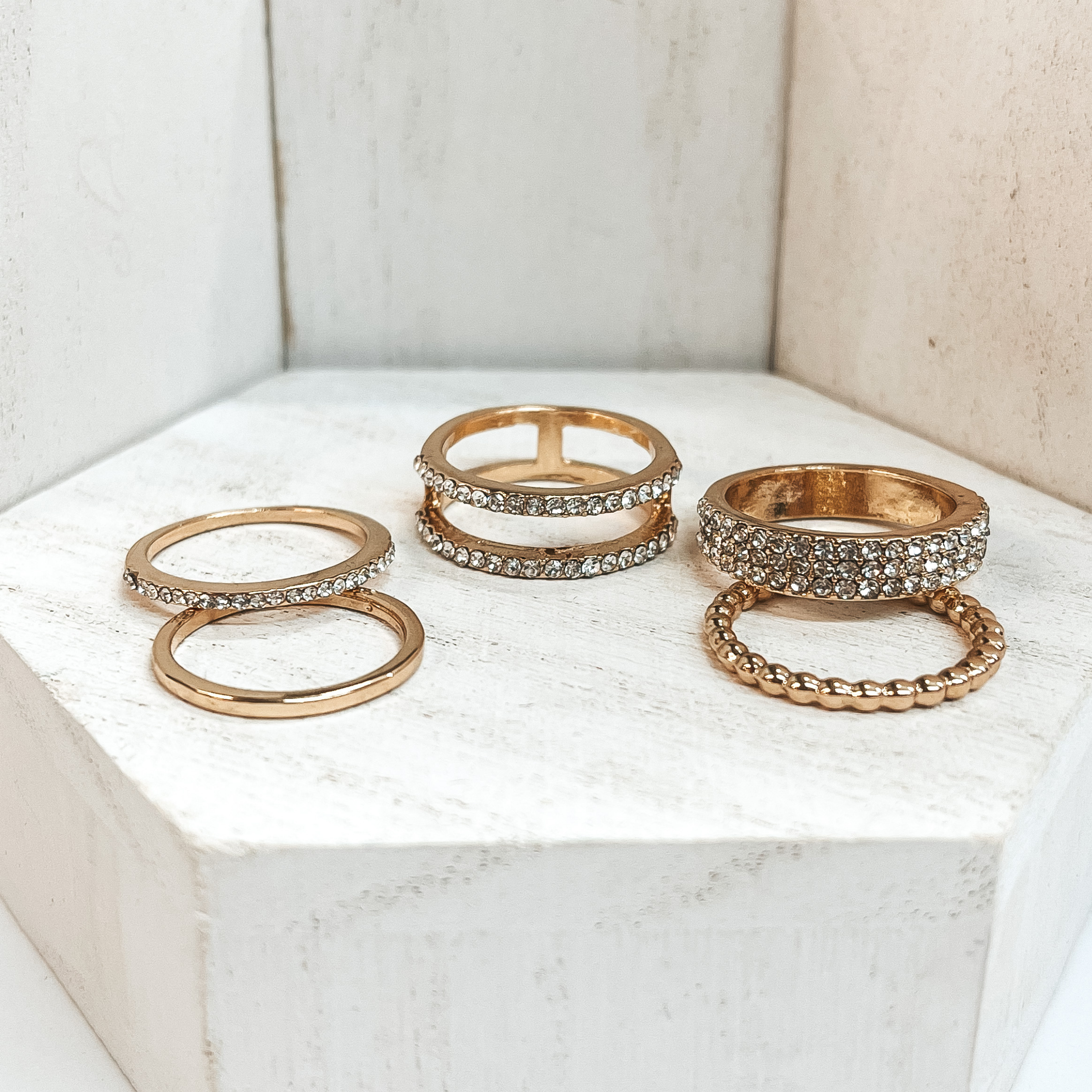 This is a set of five gold rings. All of them but  two rings have crystals in different varieties on  them. One of the rings is bubbled textured and the  other is a solid band. These rings are pictured on a white block and  white background.